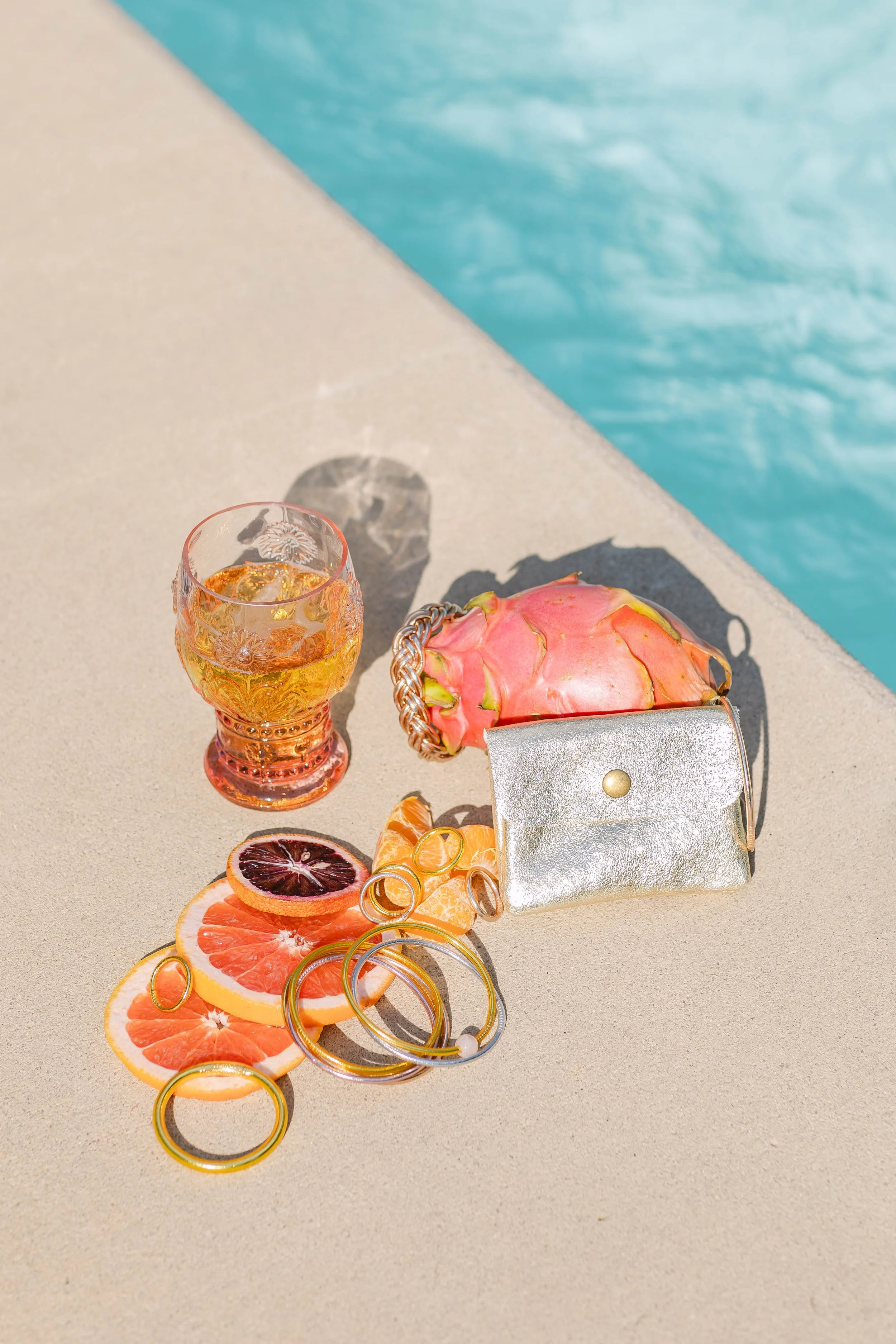 Silver mini purse, gold rings, blood orange slices, and a cocktail styled on a sun-drenched poolside surface — commercial product photography by Chandler Cox, Columbia, SC