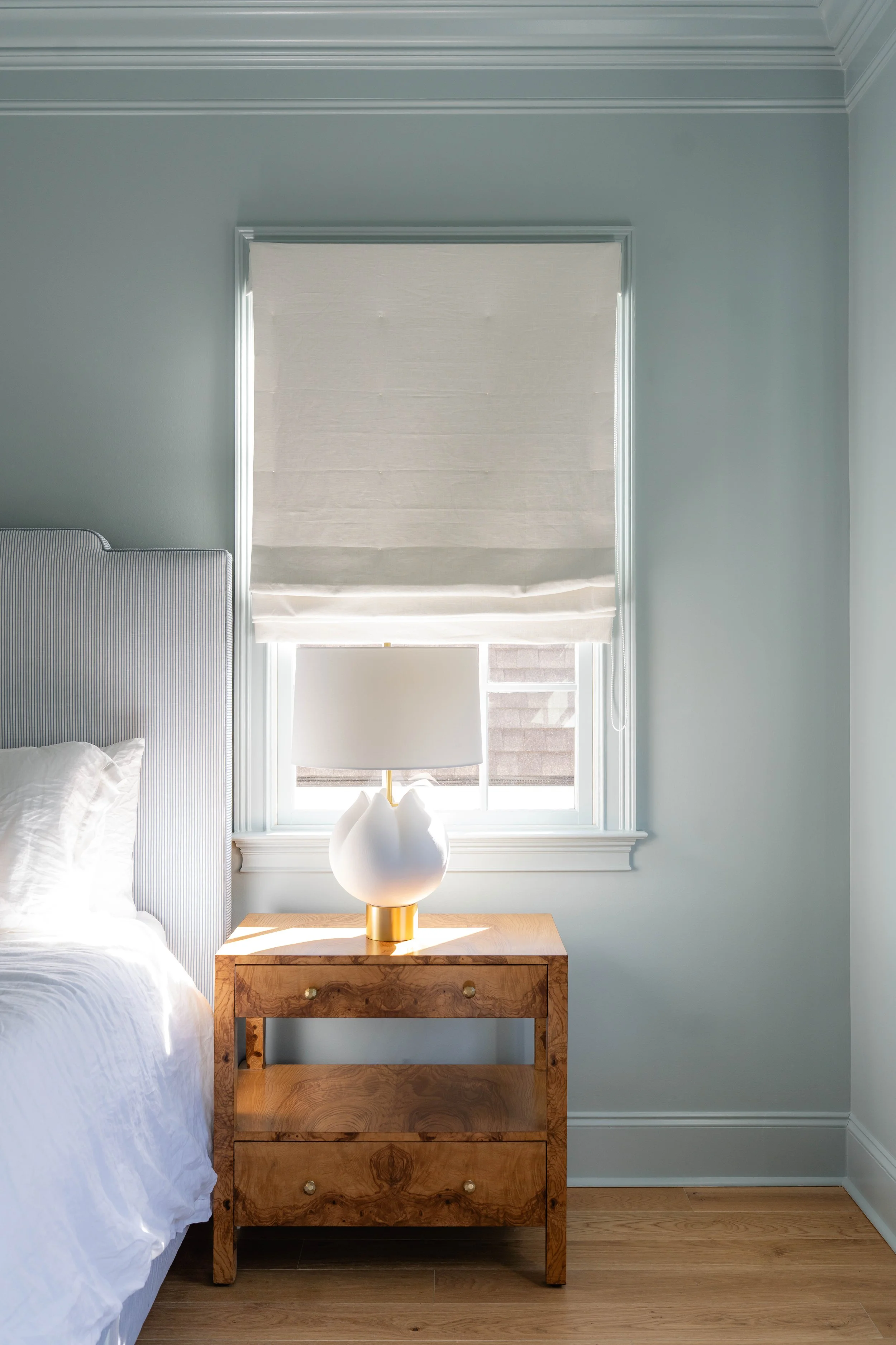Bedroom with a white bed, a wooden nightstand with two drawers, a white table lamp, a window with a white Roman shade, and light-colored walls and flooring.