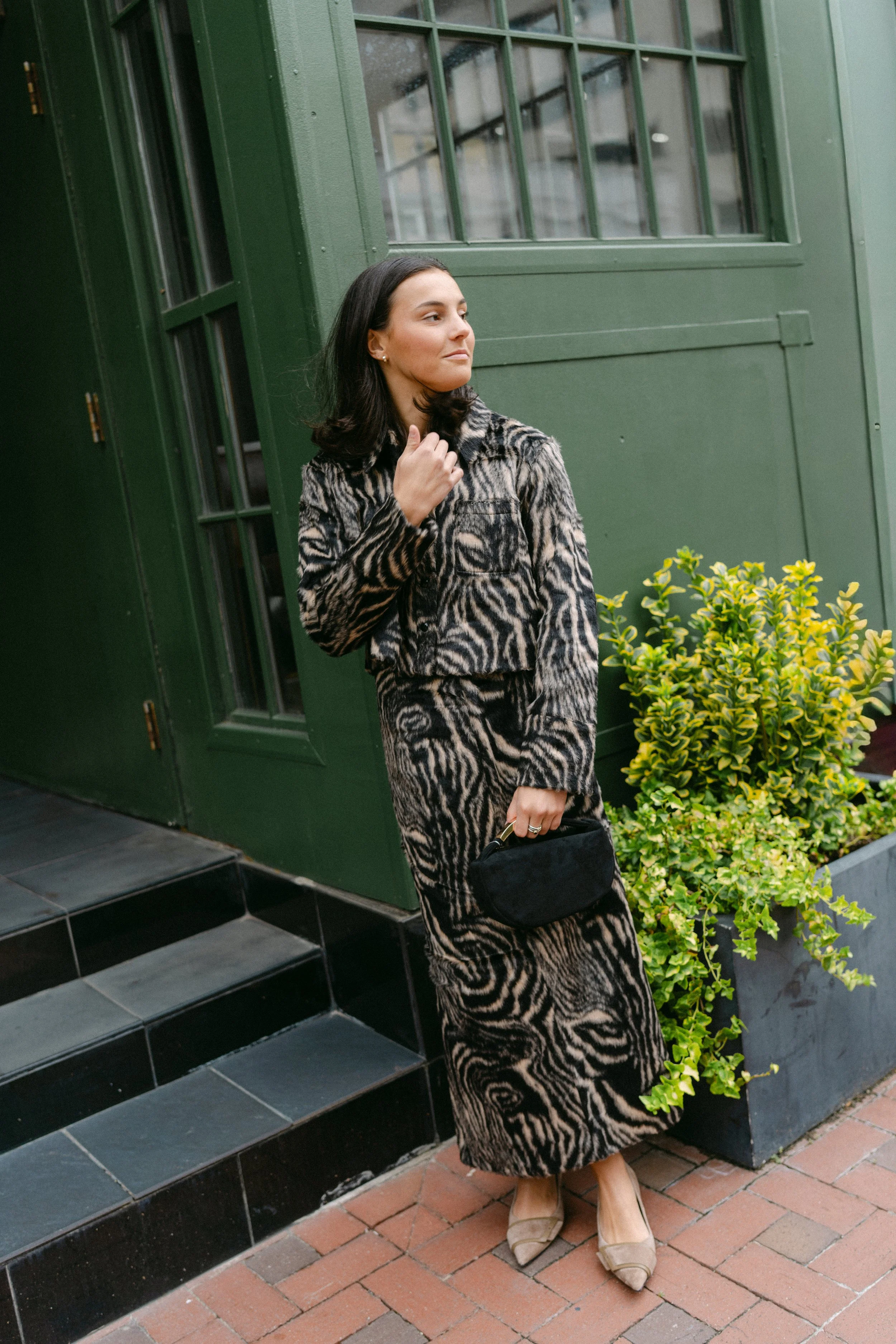 Woman in a zebra-print jacket and skirt standing beside a green storefront on a brick sidewalk during a commercial brand photography session in downtown Columbia, SC