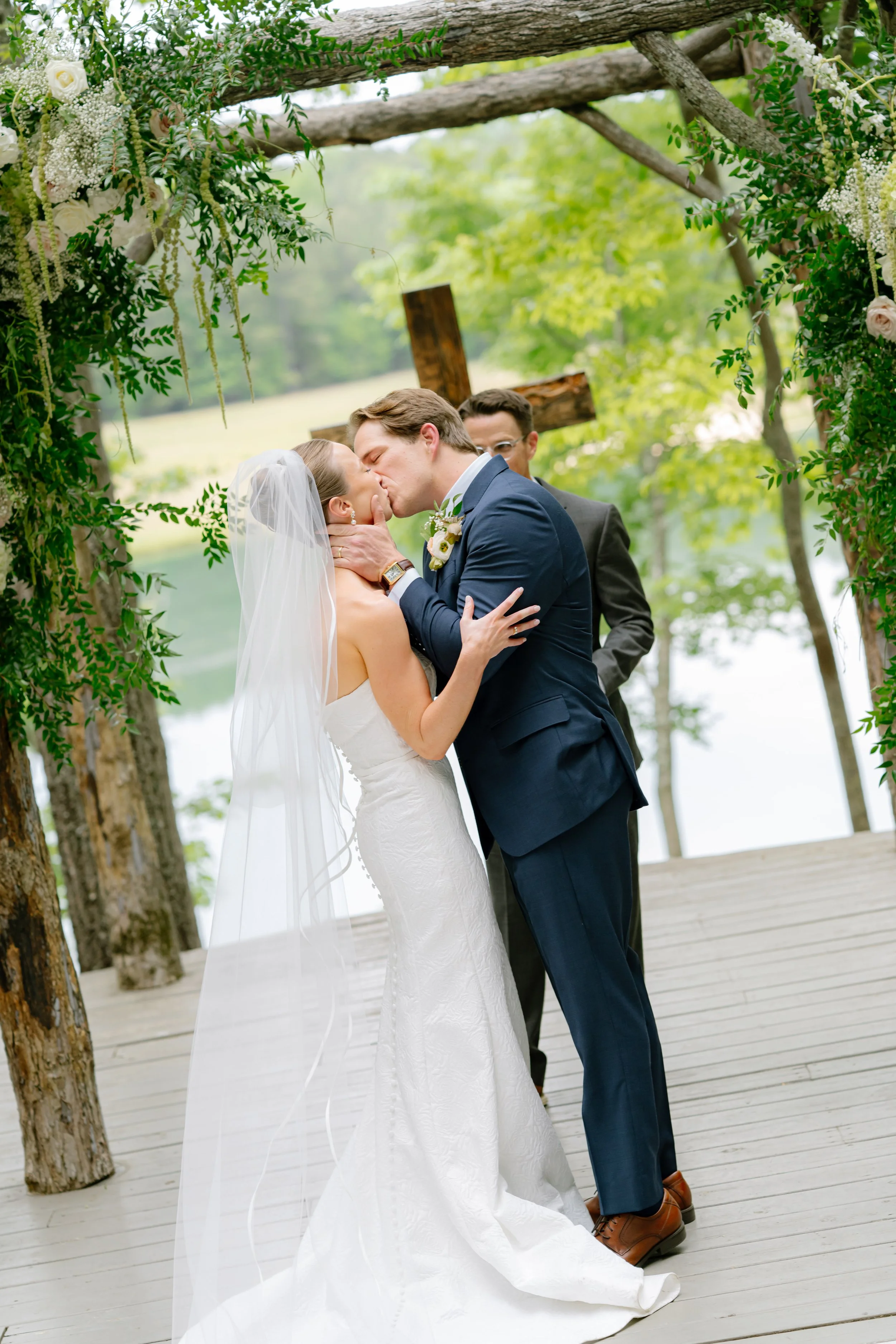 Bride and groom sharing first kiss under wooden greenery arch at lakeside wedding ceremony, South Carolina wedding photographer