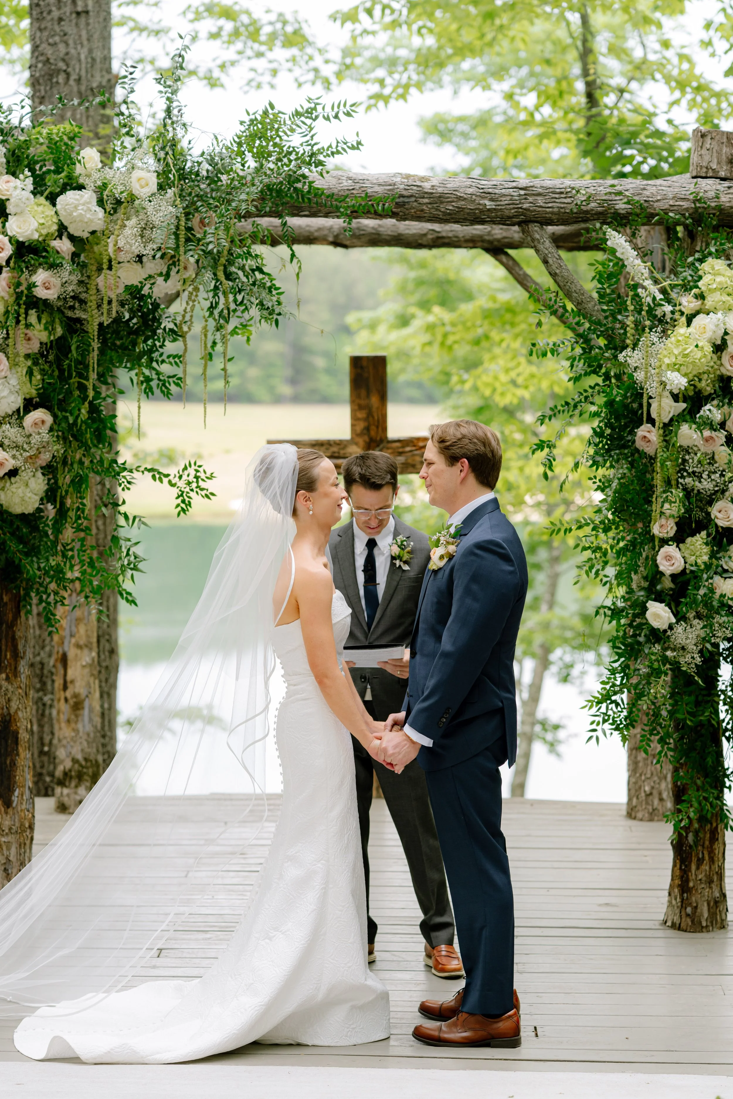 Bride and groom exchanging vows at rustic wooden arch with white florals, lakeside wedding ceremony South Carolina