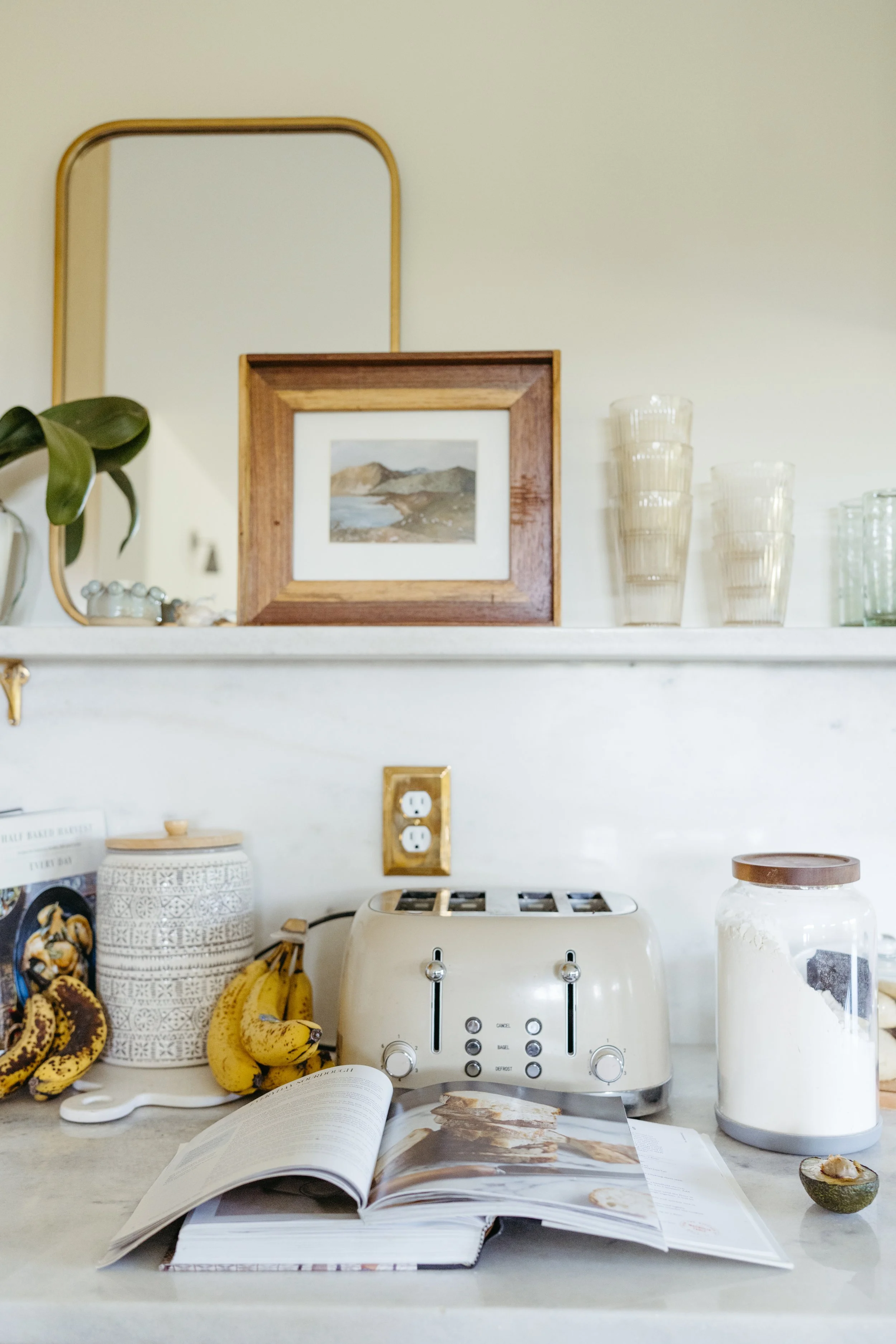 Kitchen counter with a toaster, bananas, a jar of flour, open recipe book, framed picture, and stacked glasses.