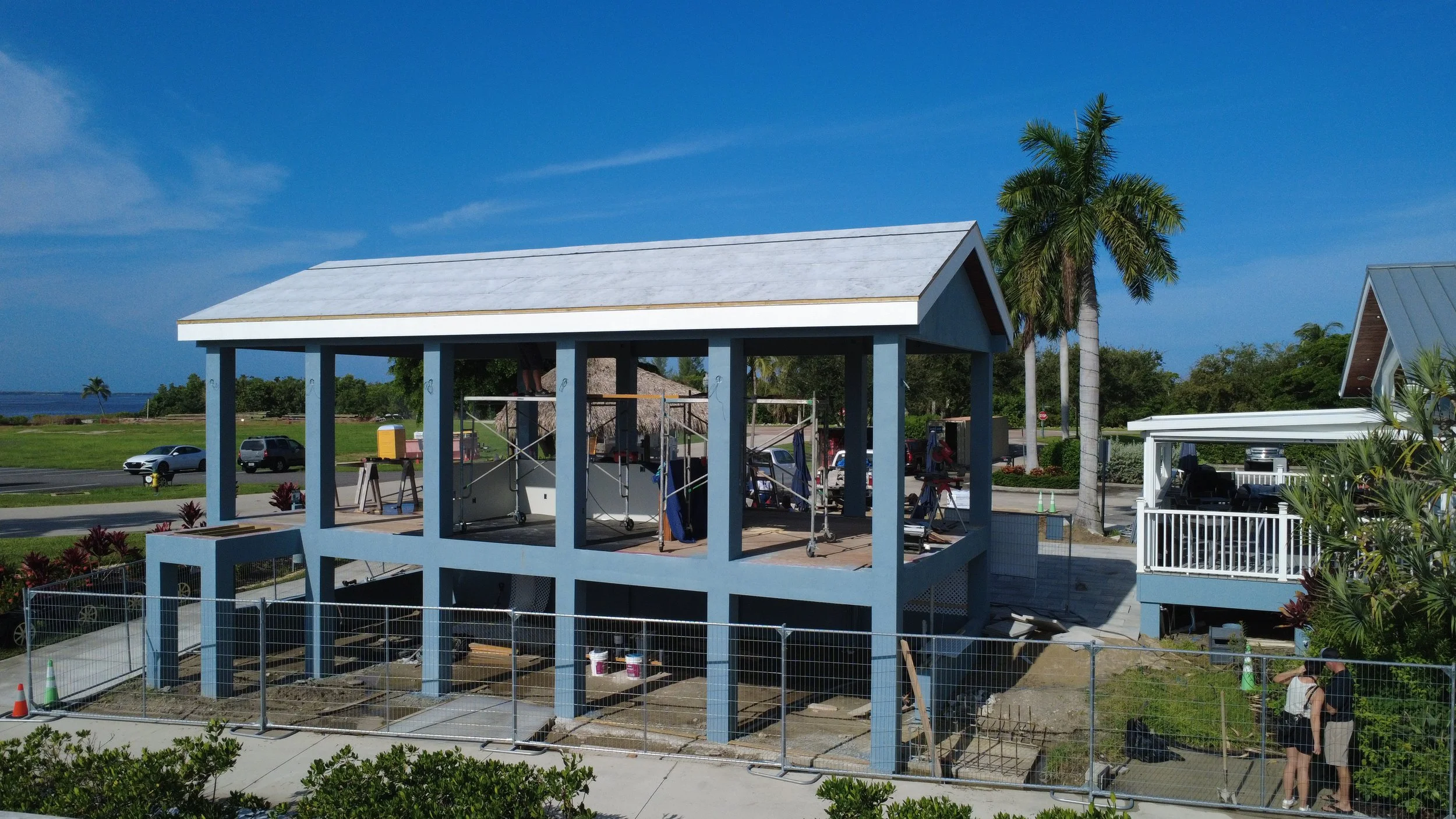 Construction site with a wooden structure being built, surrounded by a metal fence, palm trees, parked cars, and blue sky.