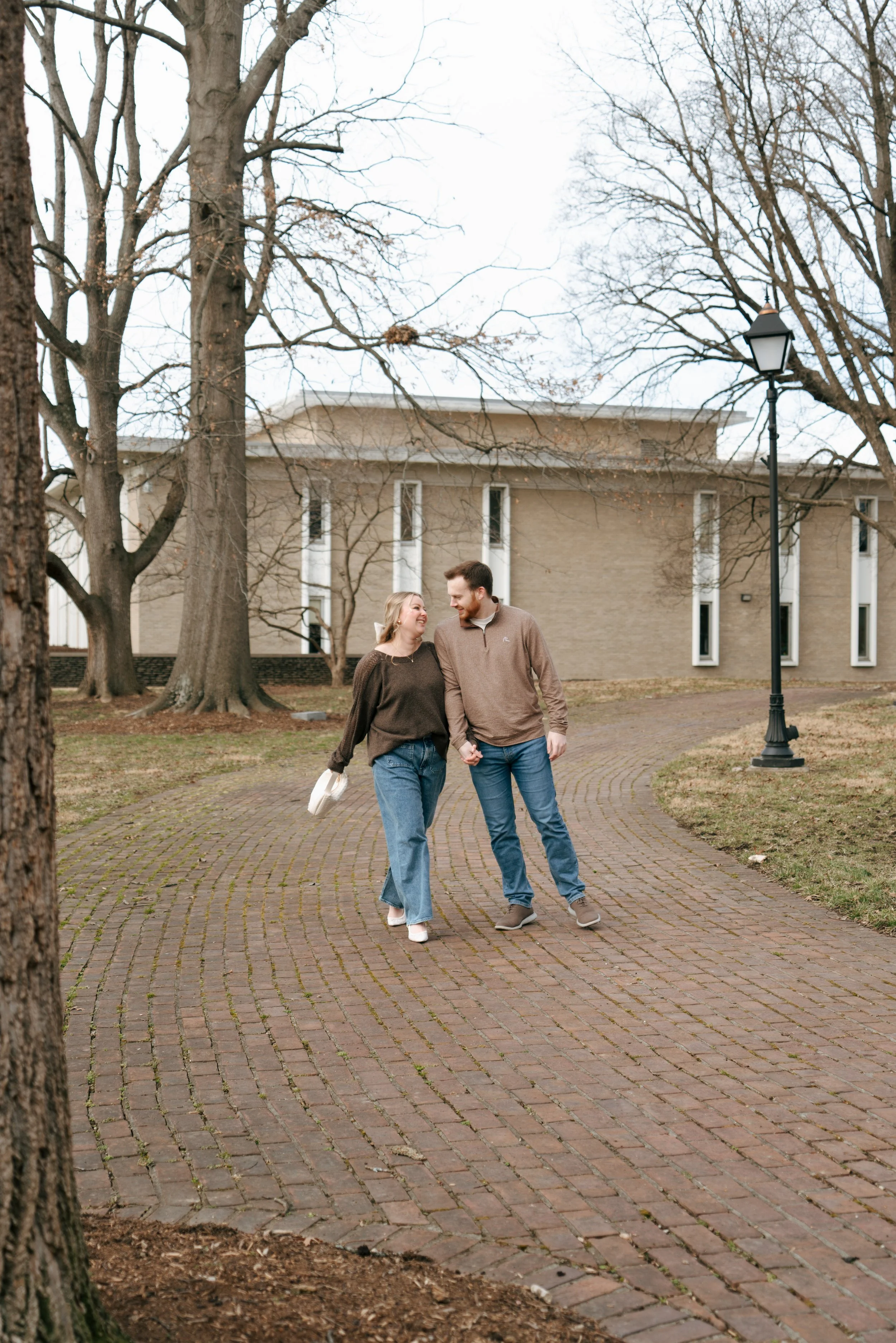 A couple holding hands and walking on a brick path in a park during fall, with leafless trees and a building in the background.