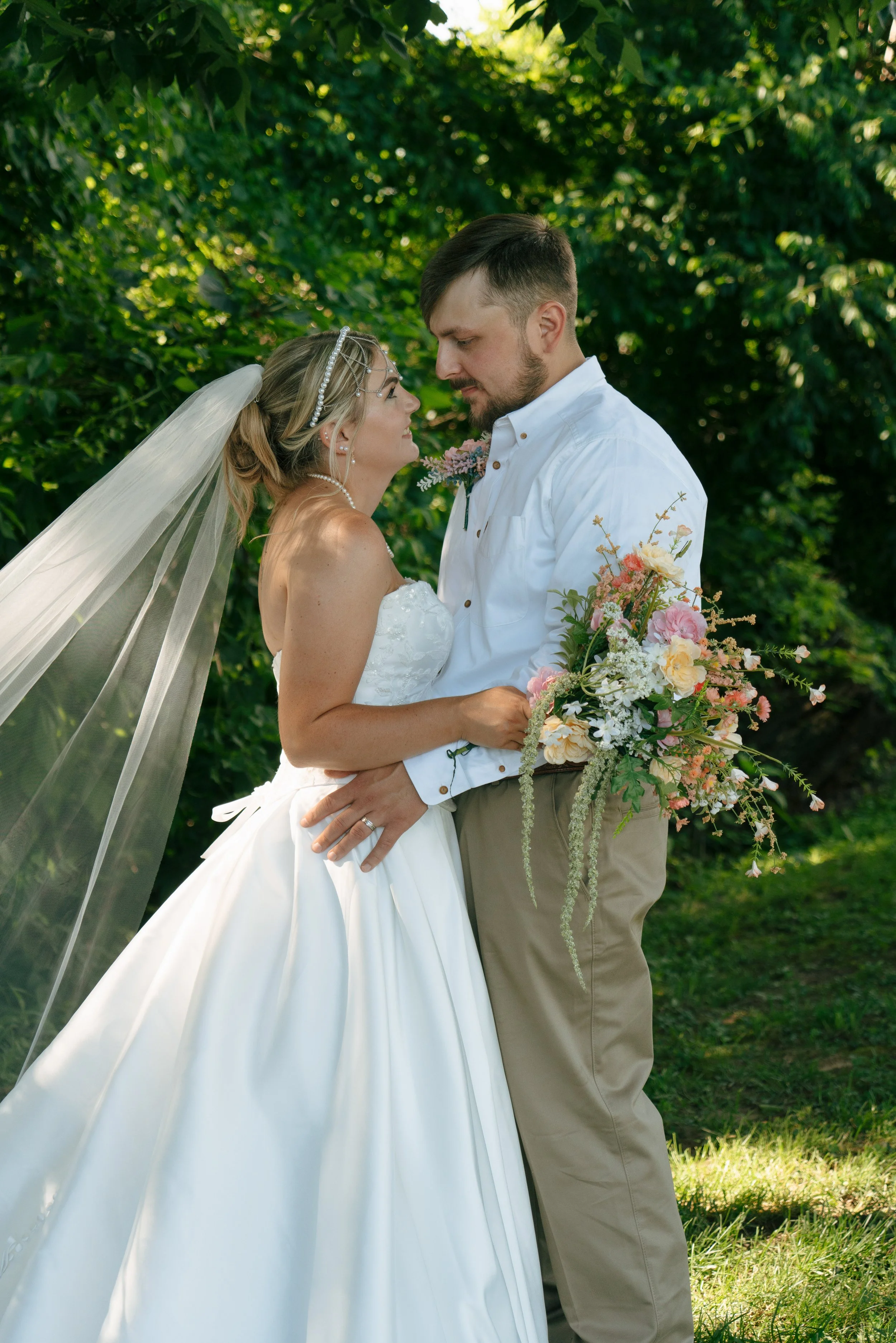 A bride and groom are standing face-to-face outdoors, holding hands, with a lush green background. The bride is wearing a white wedding gown, veil, and pearl jewelry, holding a large bouquet of pastel-colored flowers. The groom is dressed in a white 
