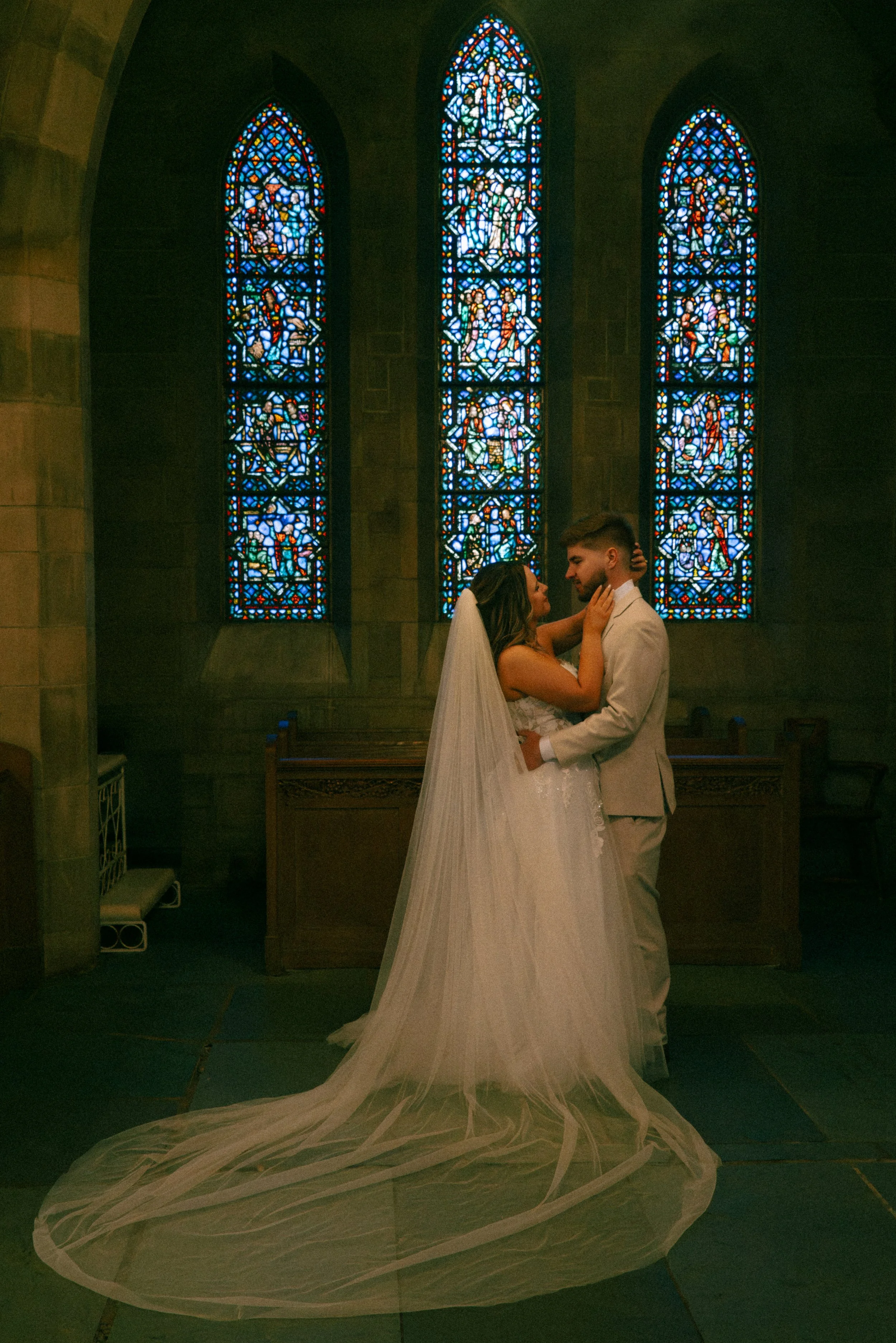 A bride and groom standing close together inside a church with stained glass windows in the background, embracing and looking into each other's eyes. The bride is wearing a wedding gown with a long train and veil, and the groom is dressed in a light-