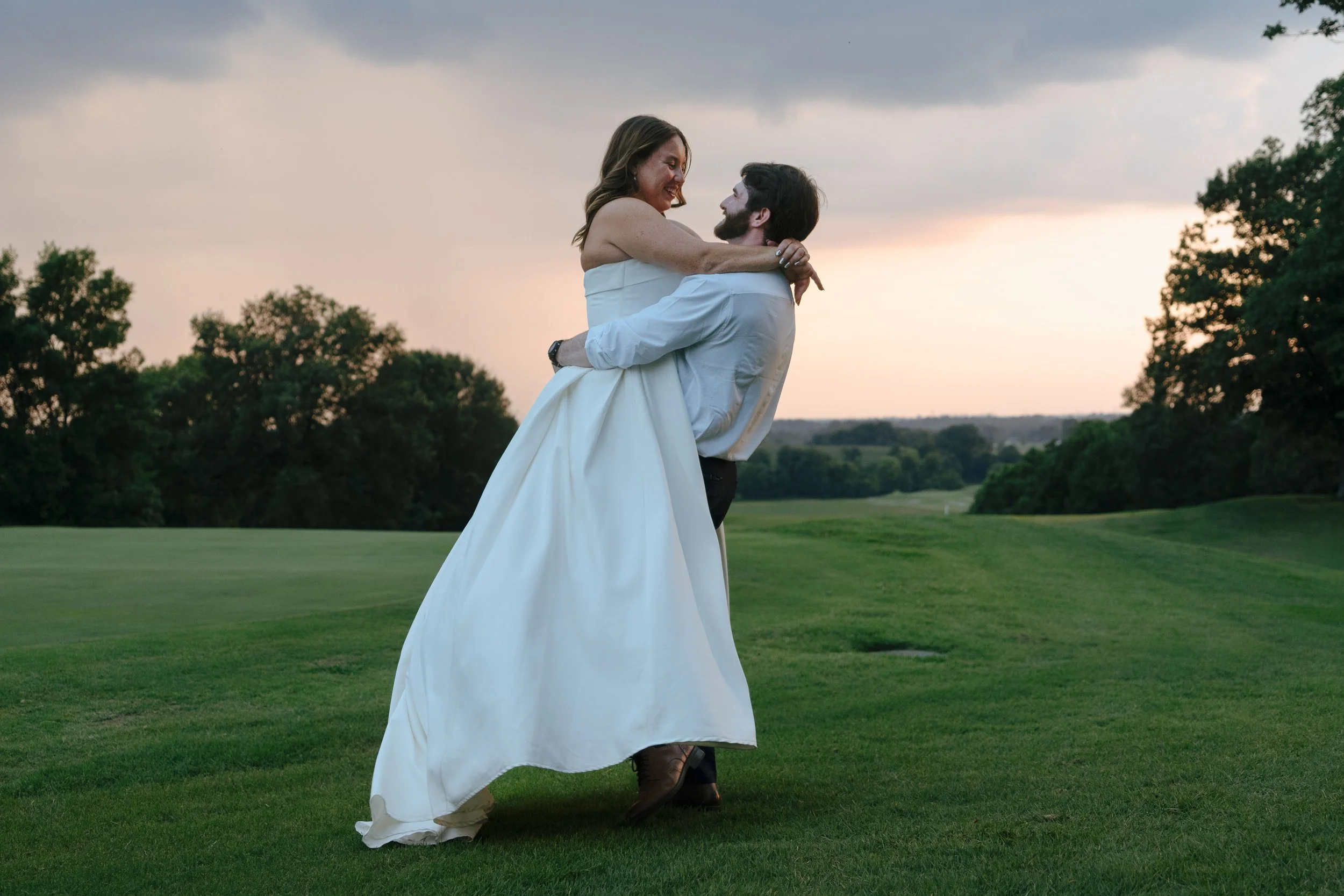 A woman in a white dress being lifted by a man in a white shirt on a grassy field during sunset, with trees in the background and a cloudy sky.