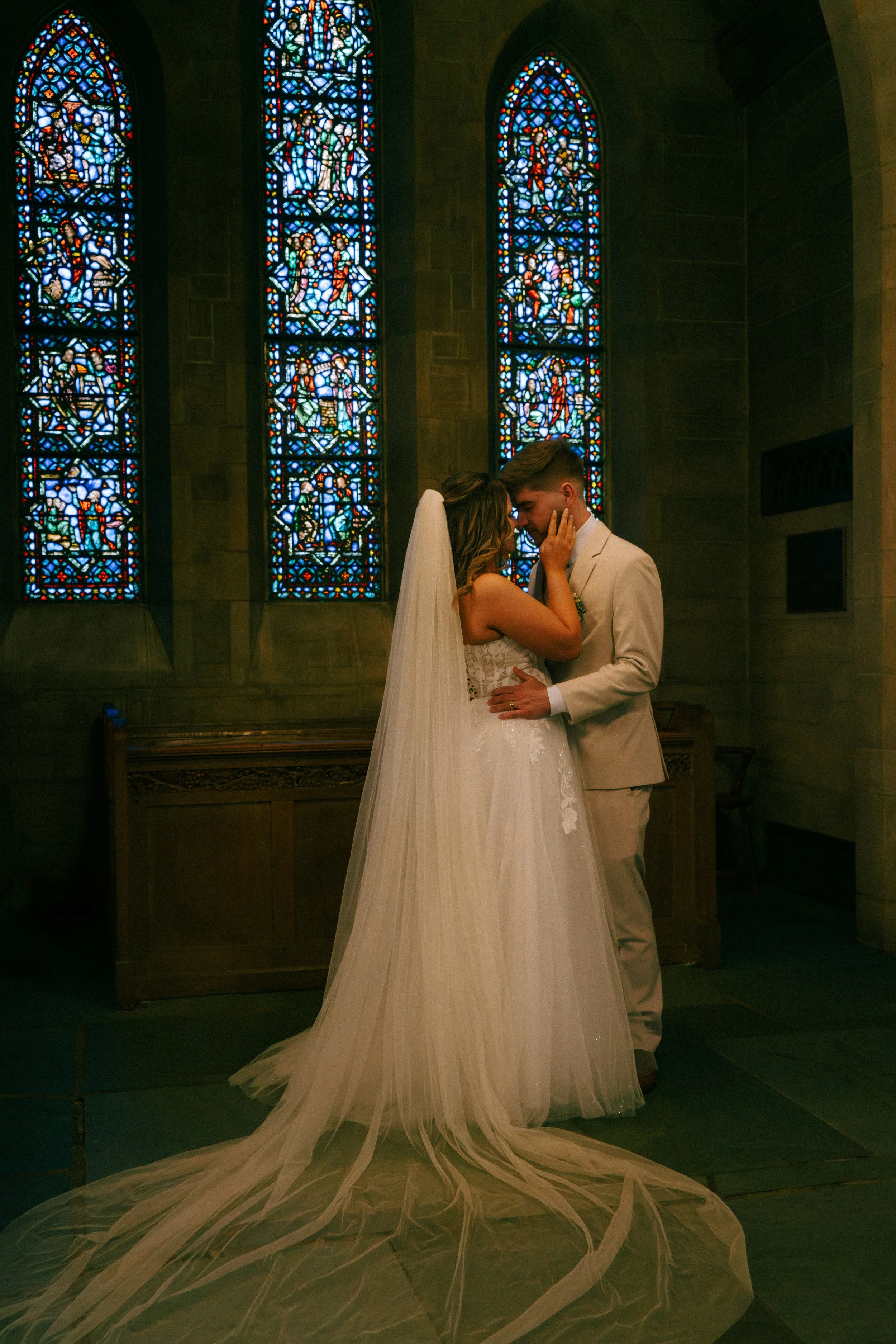 A bride and groom sharing a kiss inside a church with tall stained glass windows behind them.