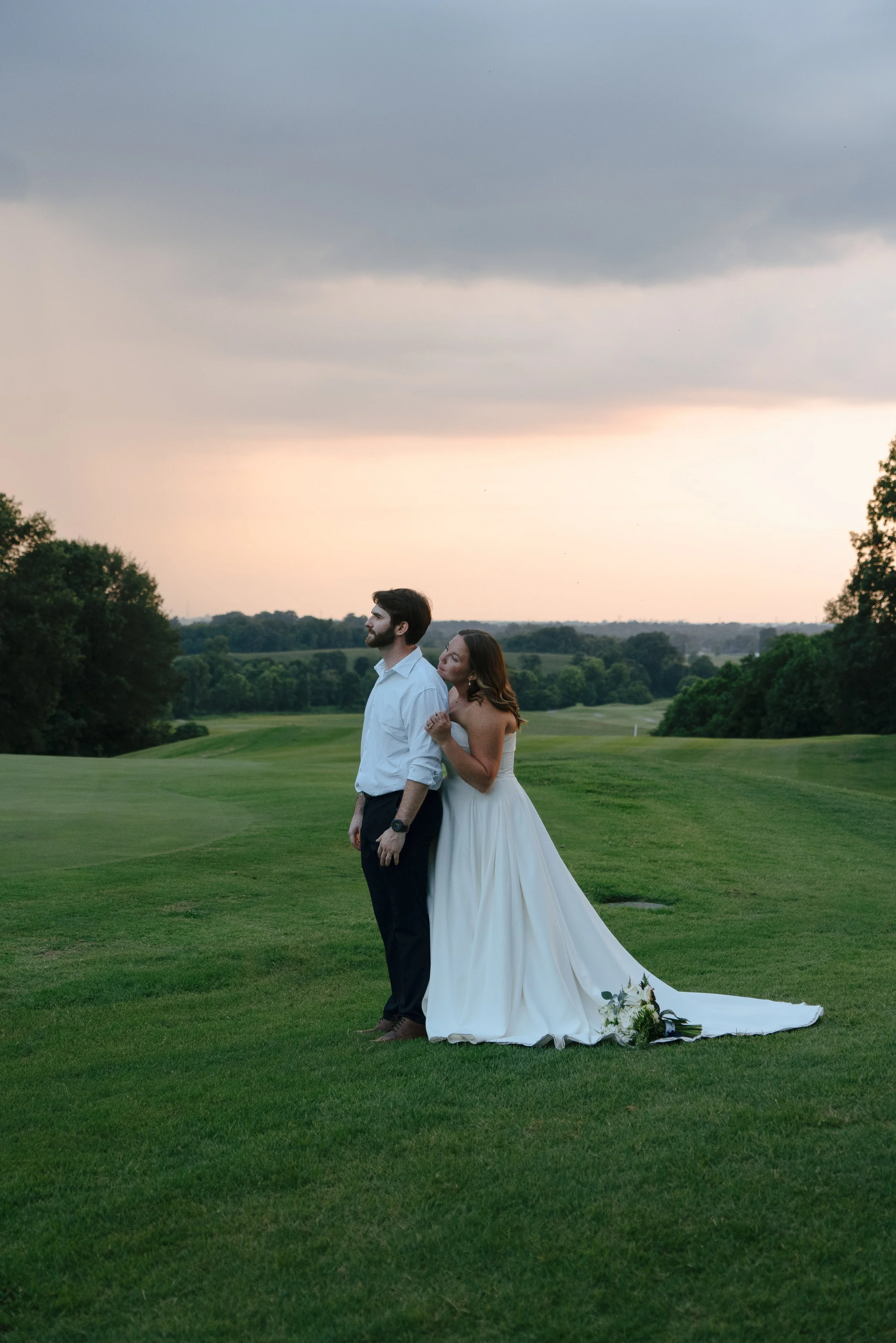 A bride and groom standing on a green golf course at sunset, with trees and a cloudy sky in the background. The bride is leaning her head on the groom's shoulder, holding his arm, and wearing a white wedding dress with a train and holding a bouquet o
