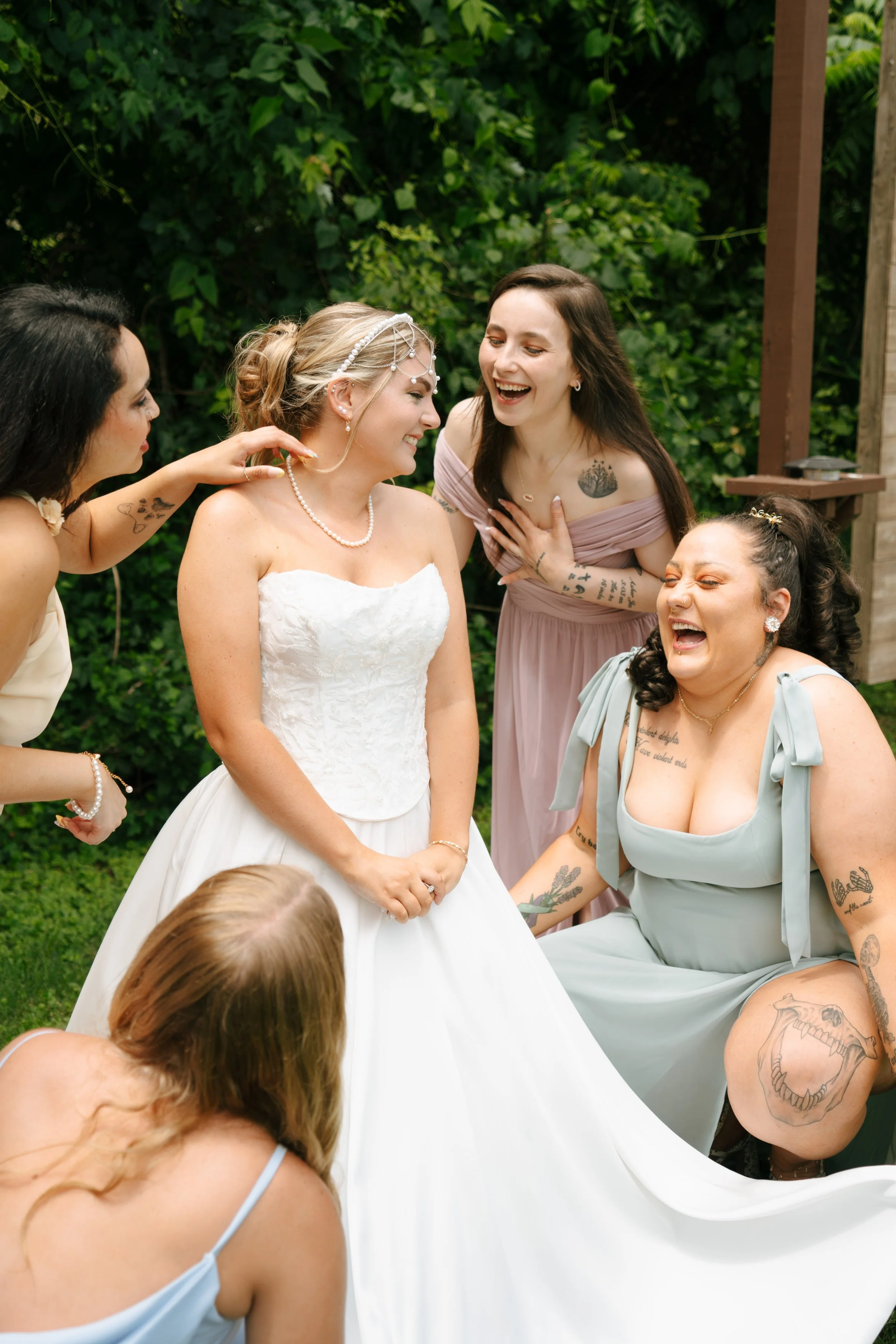 Group of women, including a bride in a white wedding dress, laughing and smiling outdoors surrounded by greenery.