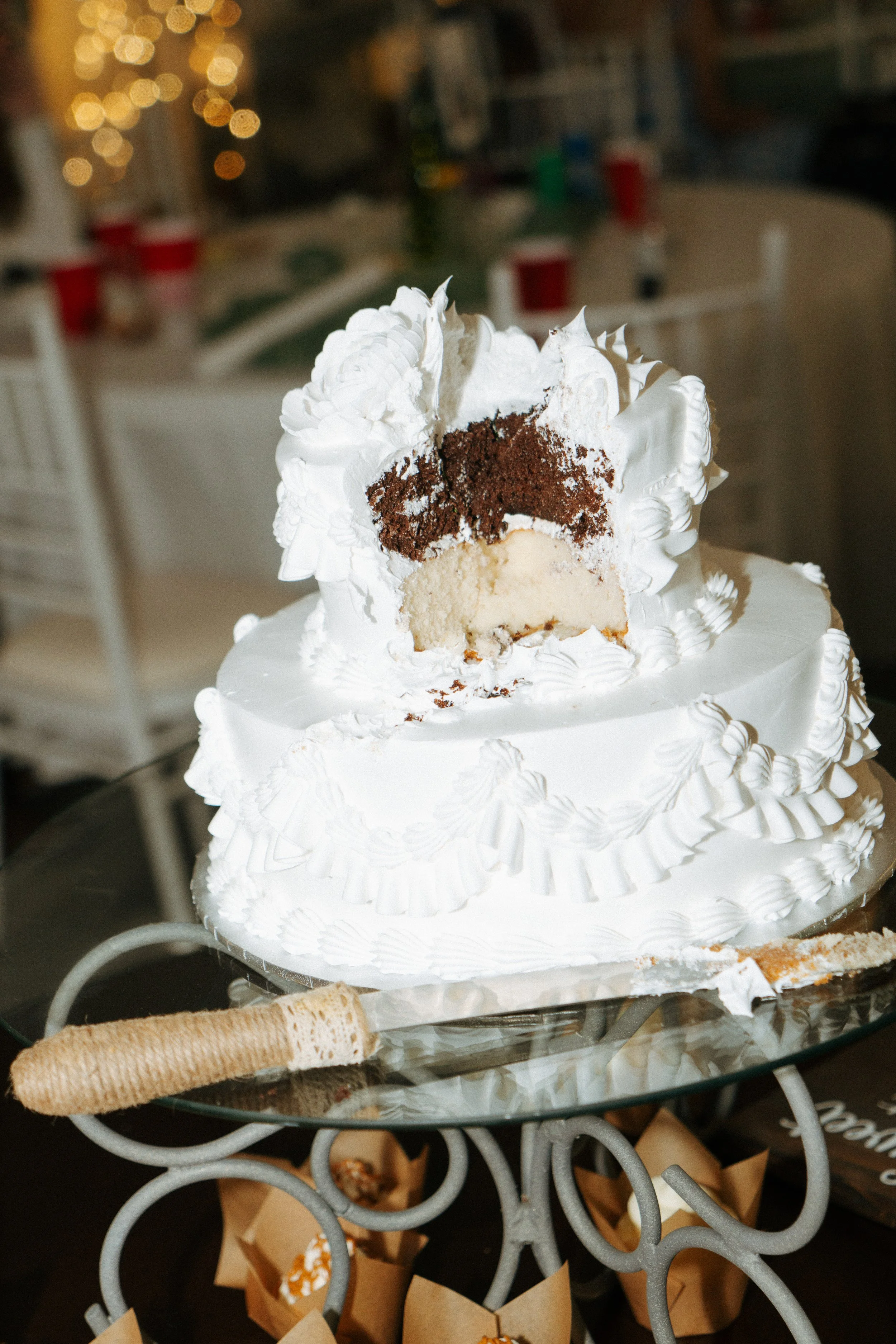 A partially sliced Christmas-themed white cake with whipped cream and chocolate layers on a decorative glass table, with blurred Christmas lights in the background.