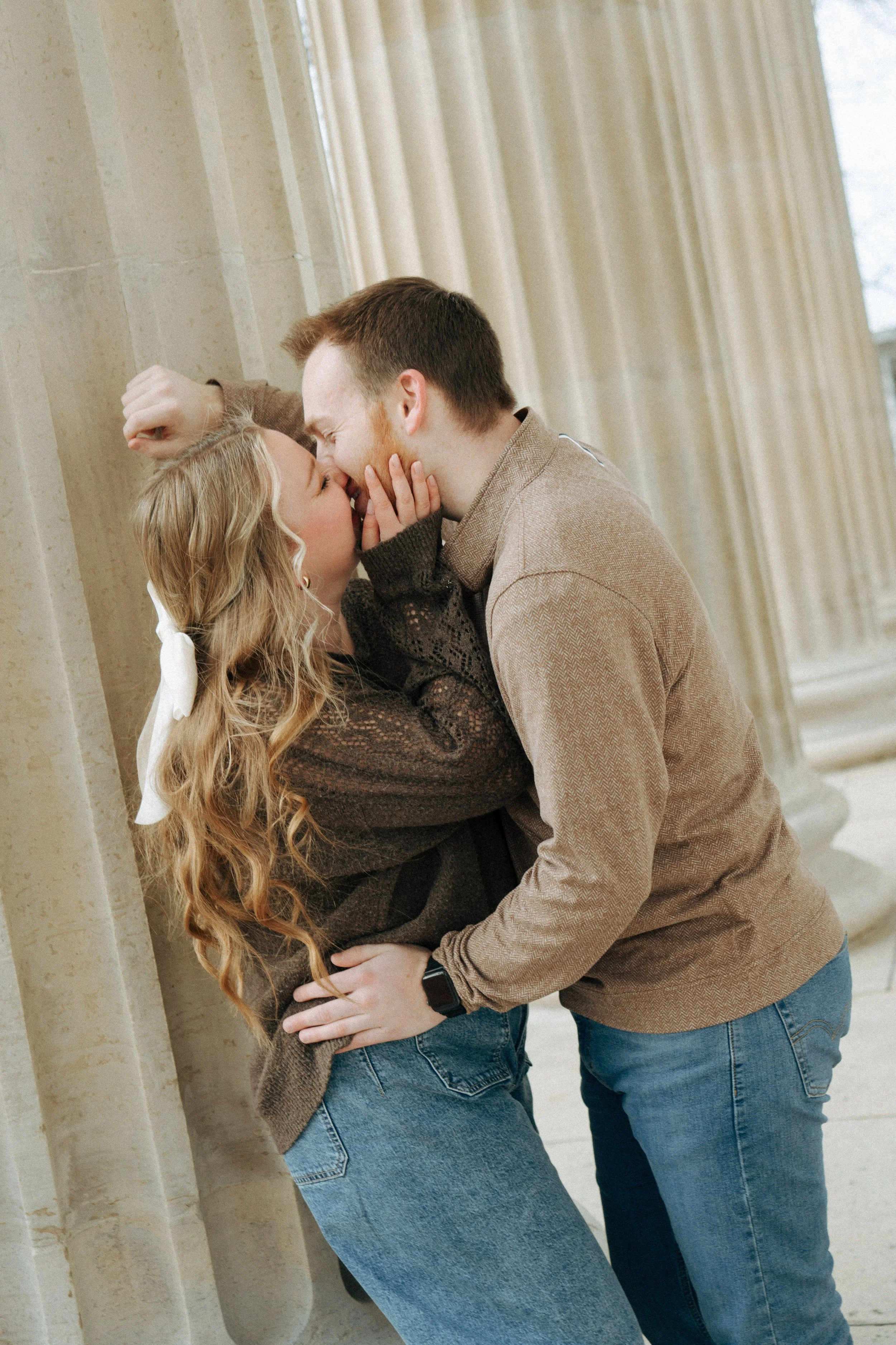 A couple kissing with their faces close together, leaning against a beige stone wall with large windows behind them.