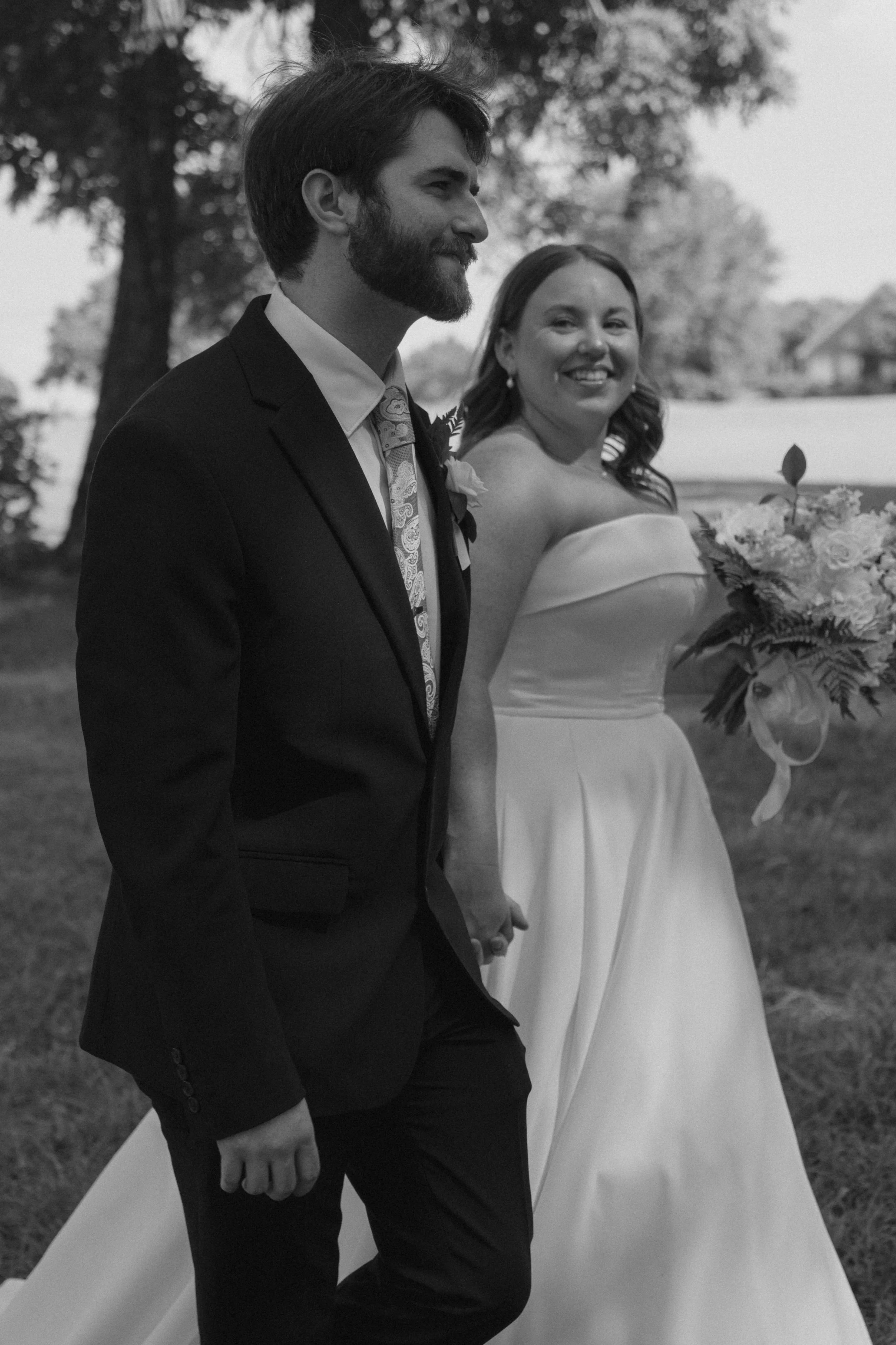 A black and white photo of a couple holding hands at their wedding outdoors. The groom, with a beard, is dressed in a suit, and the bride is smiling, holding a bouquet of flowers.
