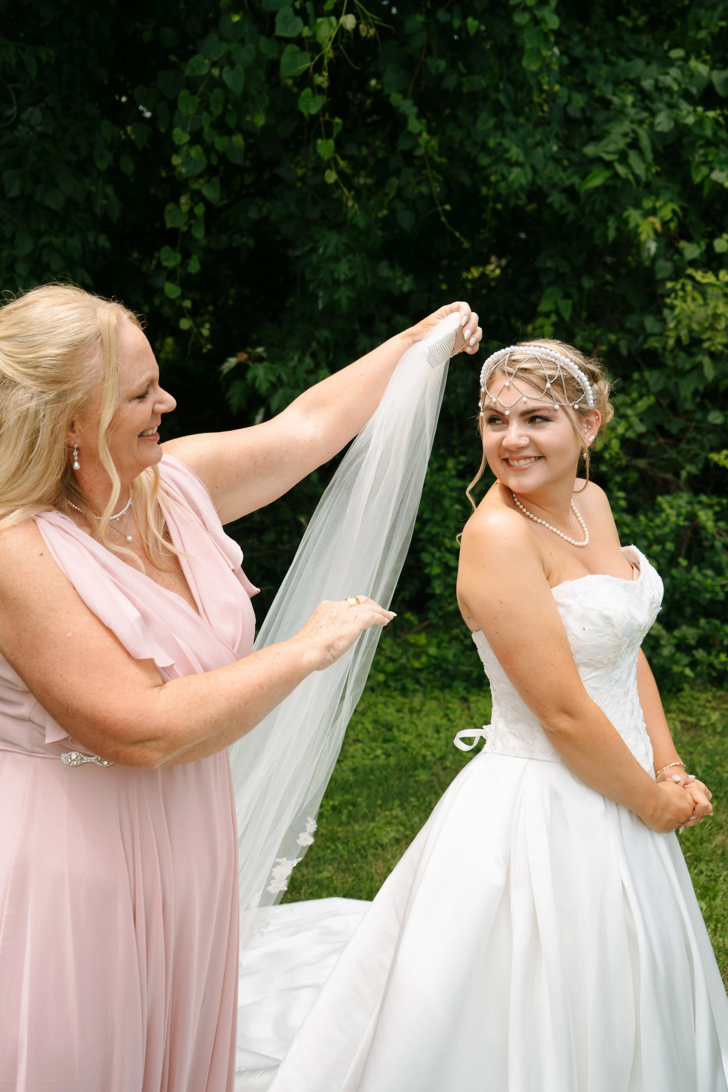 A woman in a pink dress holding a veil over a smiling bride in a white strapless wedding gown outdoors surrounded by green foliage.