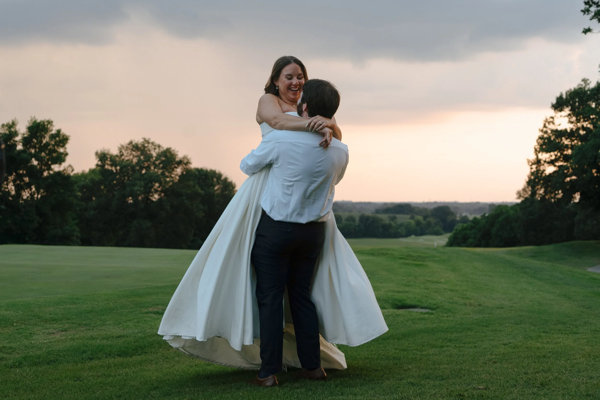 A bride and groom dancing outdoors on a grassy field during sunset with trees in the background.