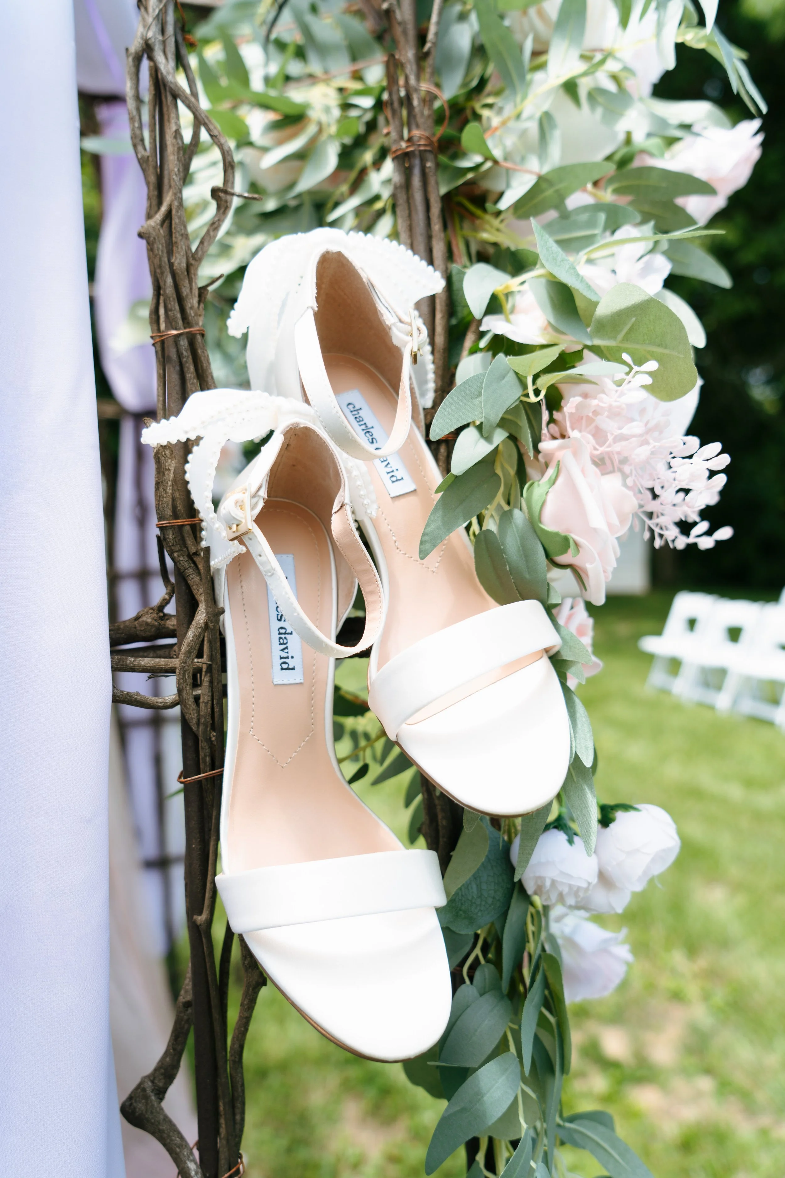 White high-heeled shoes with ankle straps hanging on a wooden structure decorated with green leaves and pale pink flowers in an outdoor setting.
