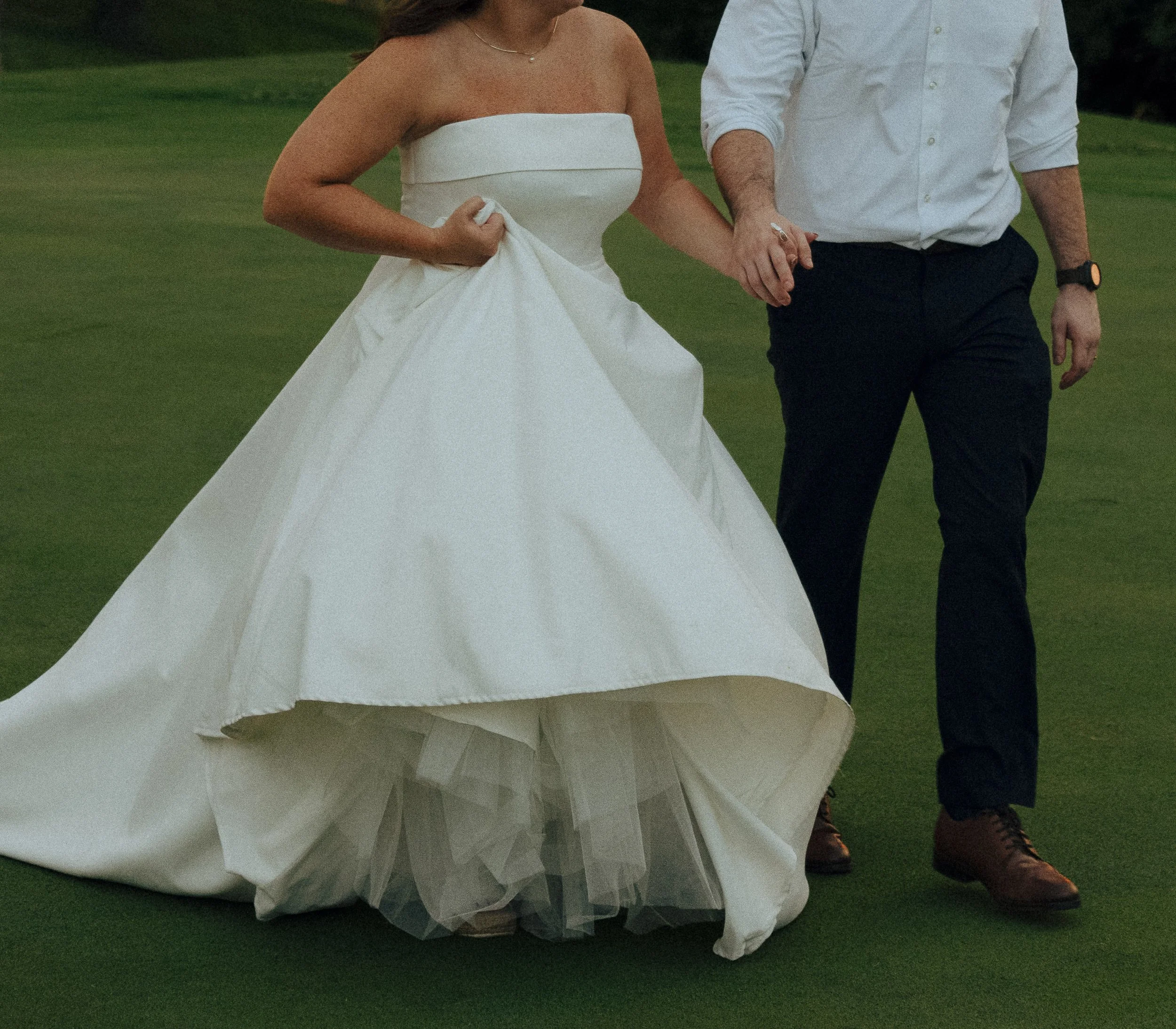 A bride in a strapless white wedding dress holding hands with a man in a white shirt and dark pants, walking on grass outdoors.