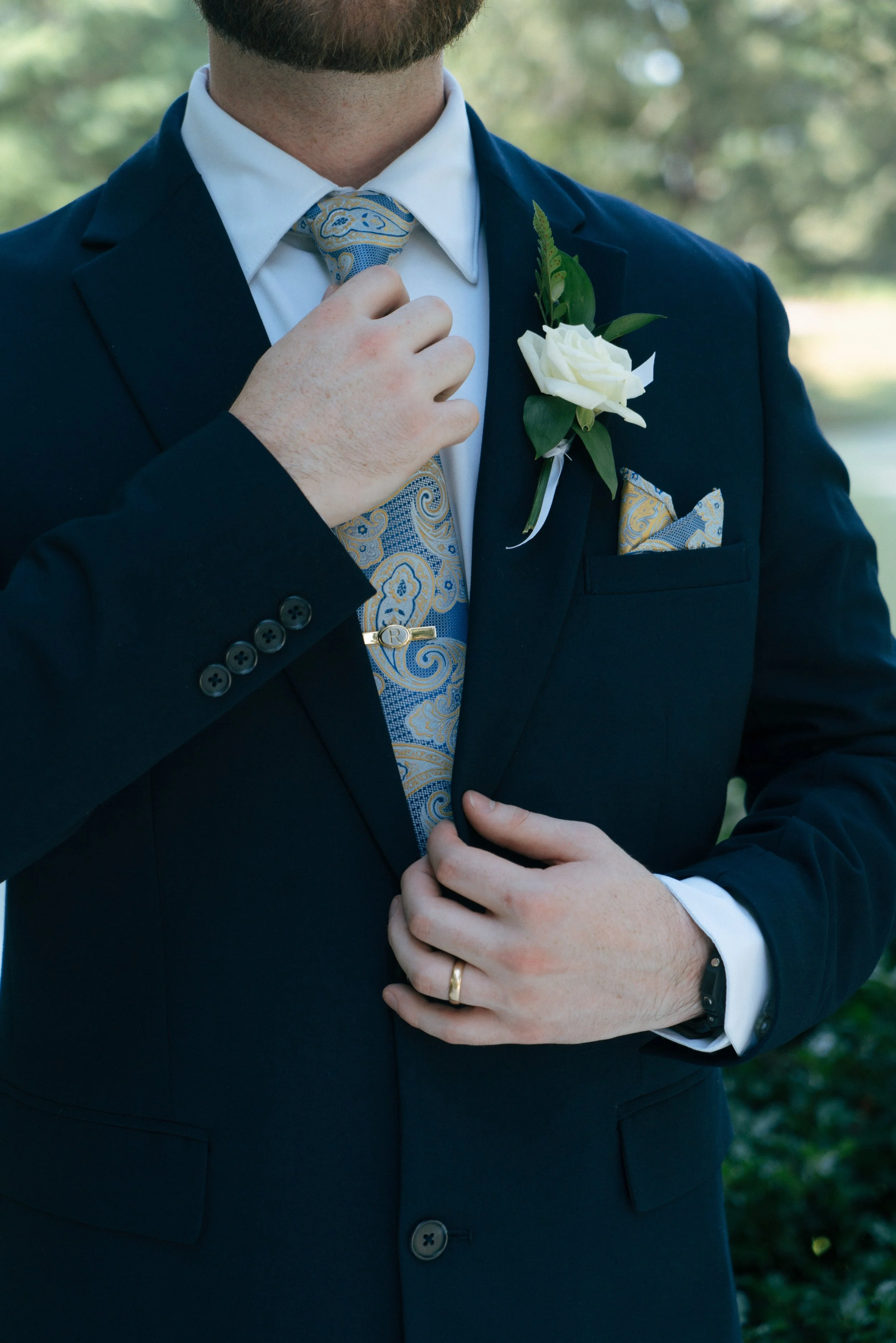 Close-up of a groom adjusting his tie. He is wearing a navy blue suit with a white dress shirt, a blue and gold patterned tie, a boutonniere with white flowers, and a pocket square. There are trees in the background.