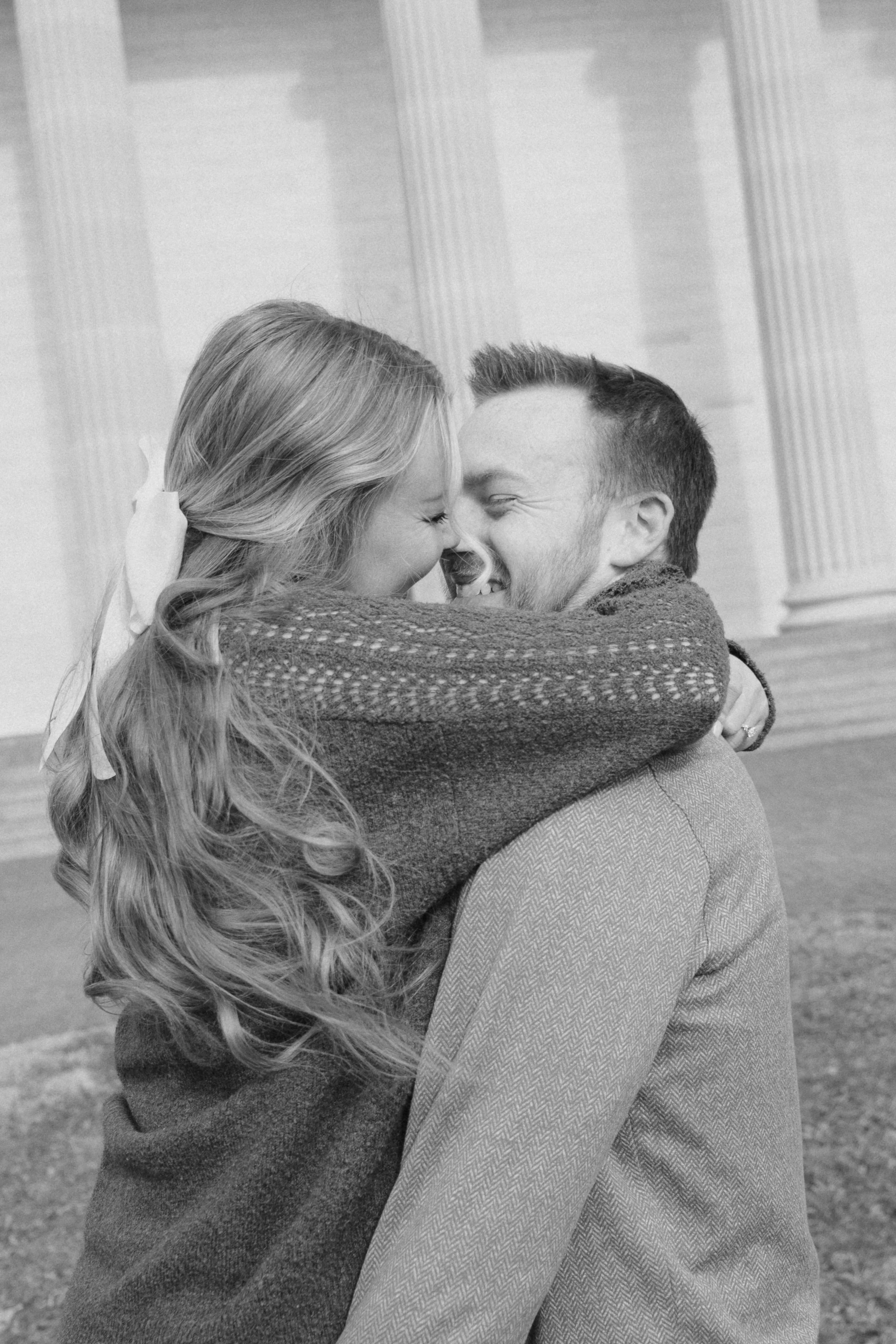 A black and white photograph of a couple embracing and smiling with their foreheads touching, outdoors near a building with columns.