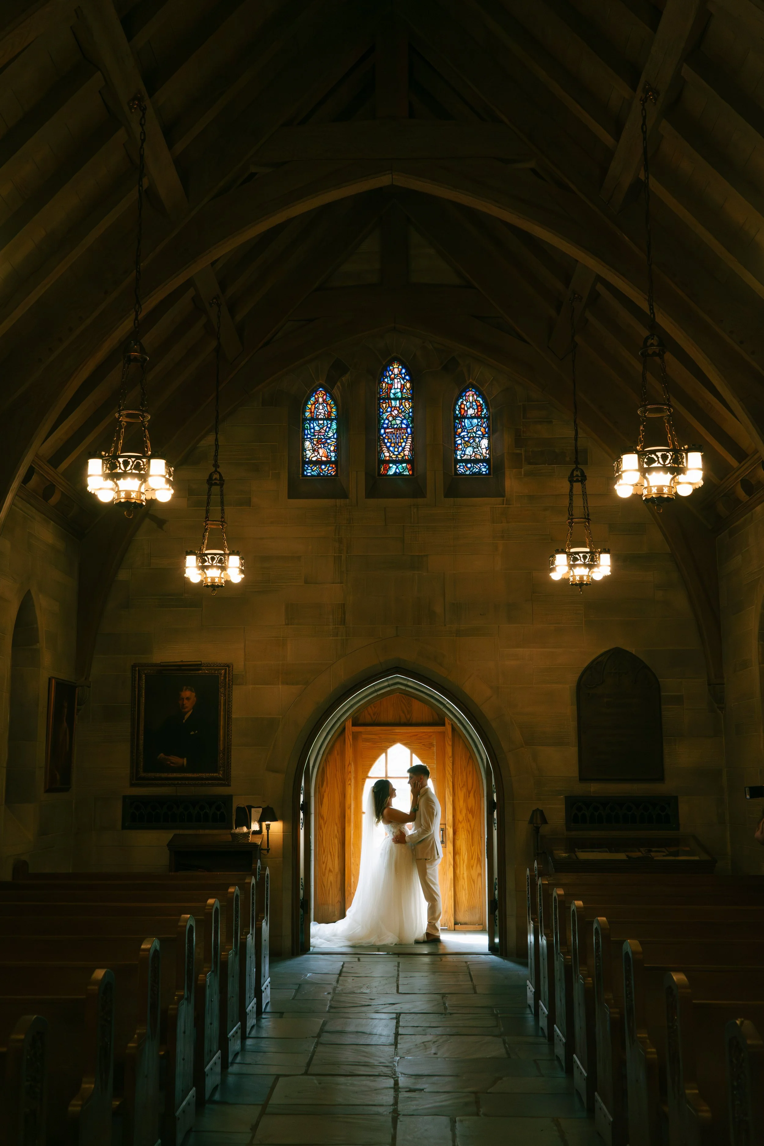 A bride and groom standing in a church doorway, holding hands and facing each other. The interior features high wooden ceilings, stained glass windows, and hanging chandeliers.