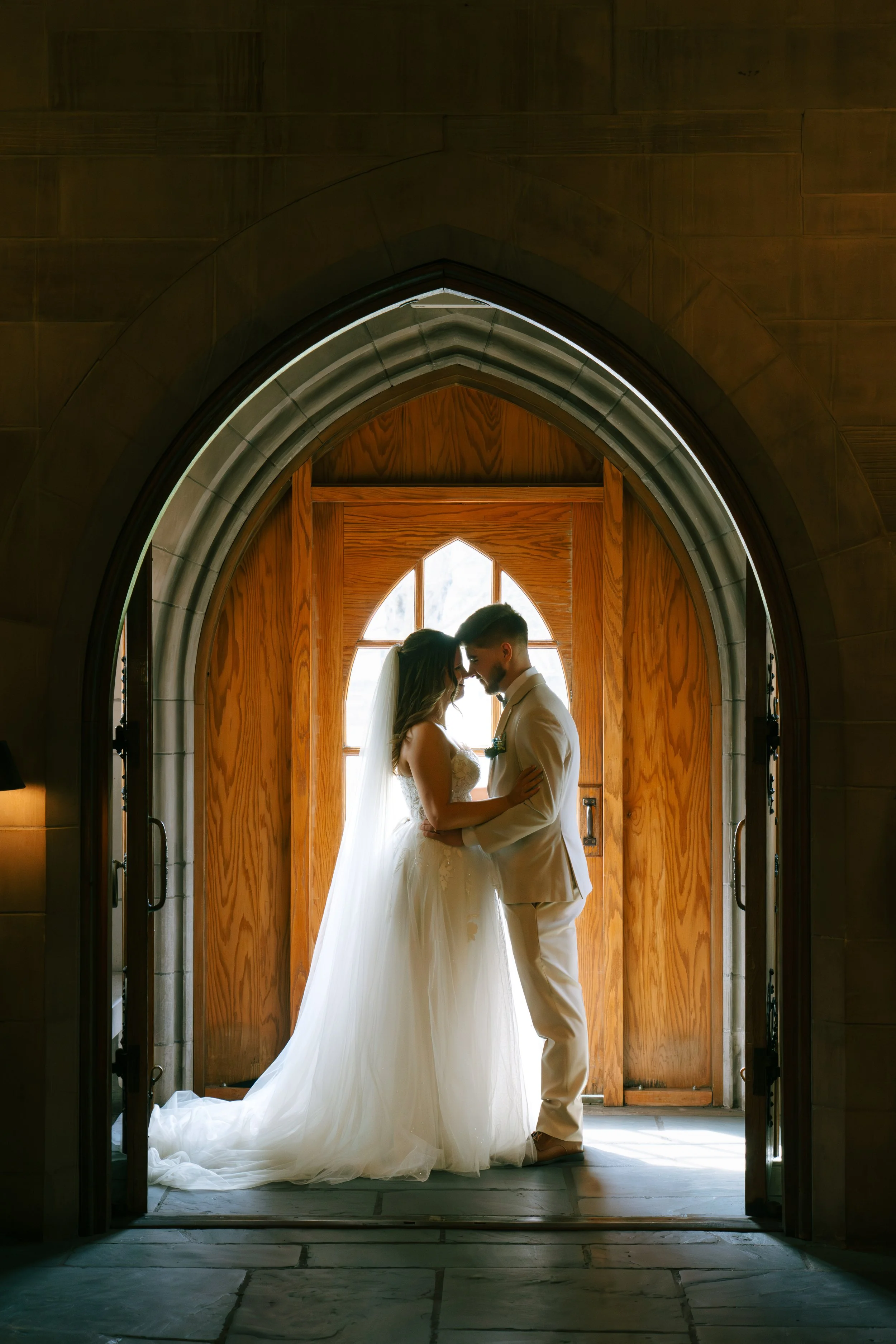 A bride and groom standing close together inside a church or cathedral archway, with the bride in a white wedding gown and veil, and the groom in a white suit, touching foreheads. Bright natural light shines through a window behind them, illuminating