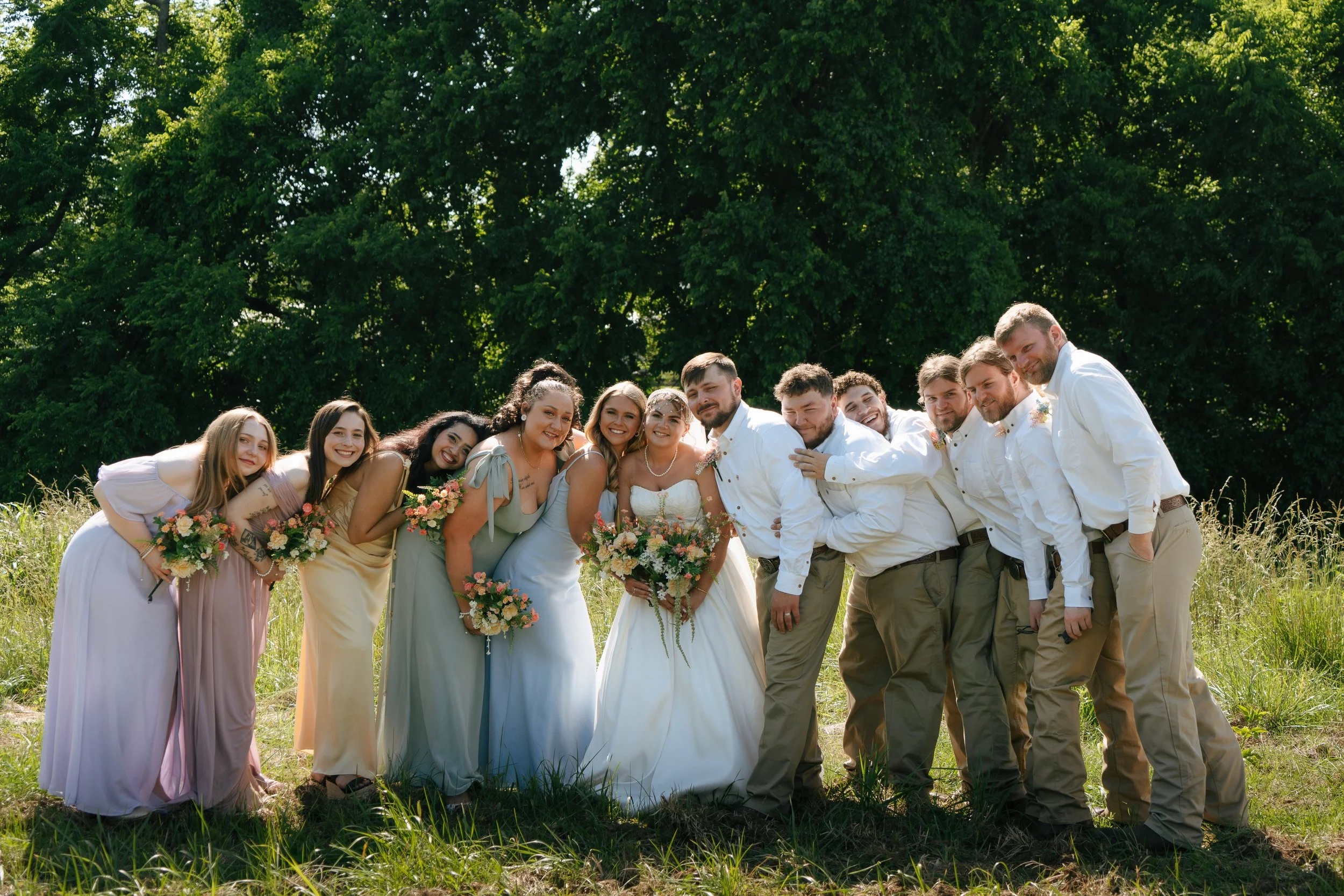 A group of people dressed in wedding attire, standing outdoors in front of green trees, smiling and posing for a photo. The women are holding bouquets, and the men are wearing white shirts and beige pants.