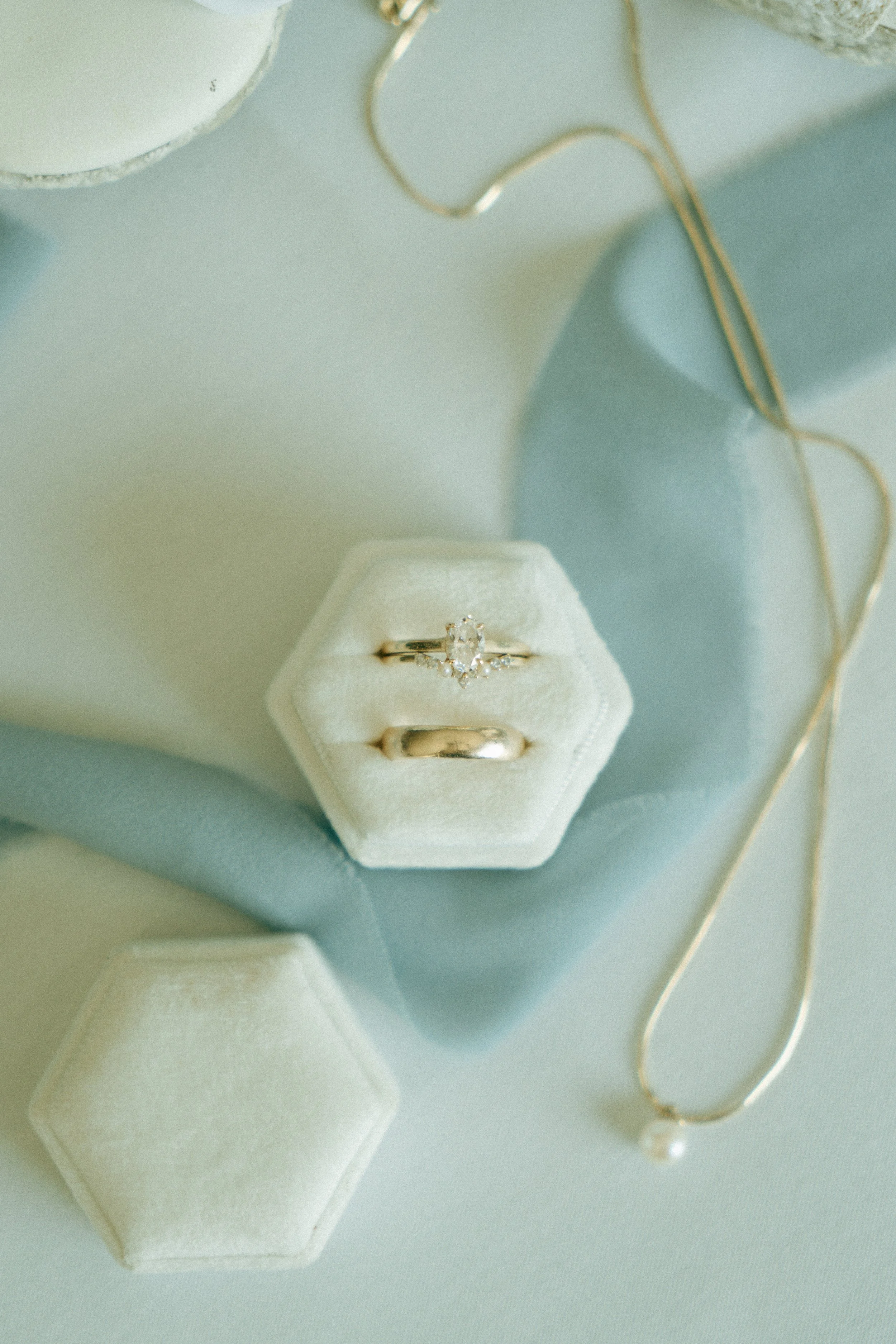Close-up of two rings in a cream-colored ring box, including a diamond engagement ring and a plain wedding band, with a gold necklace visible in the background, on a light-colored surface.