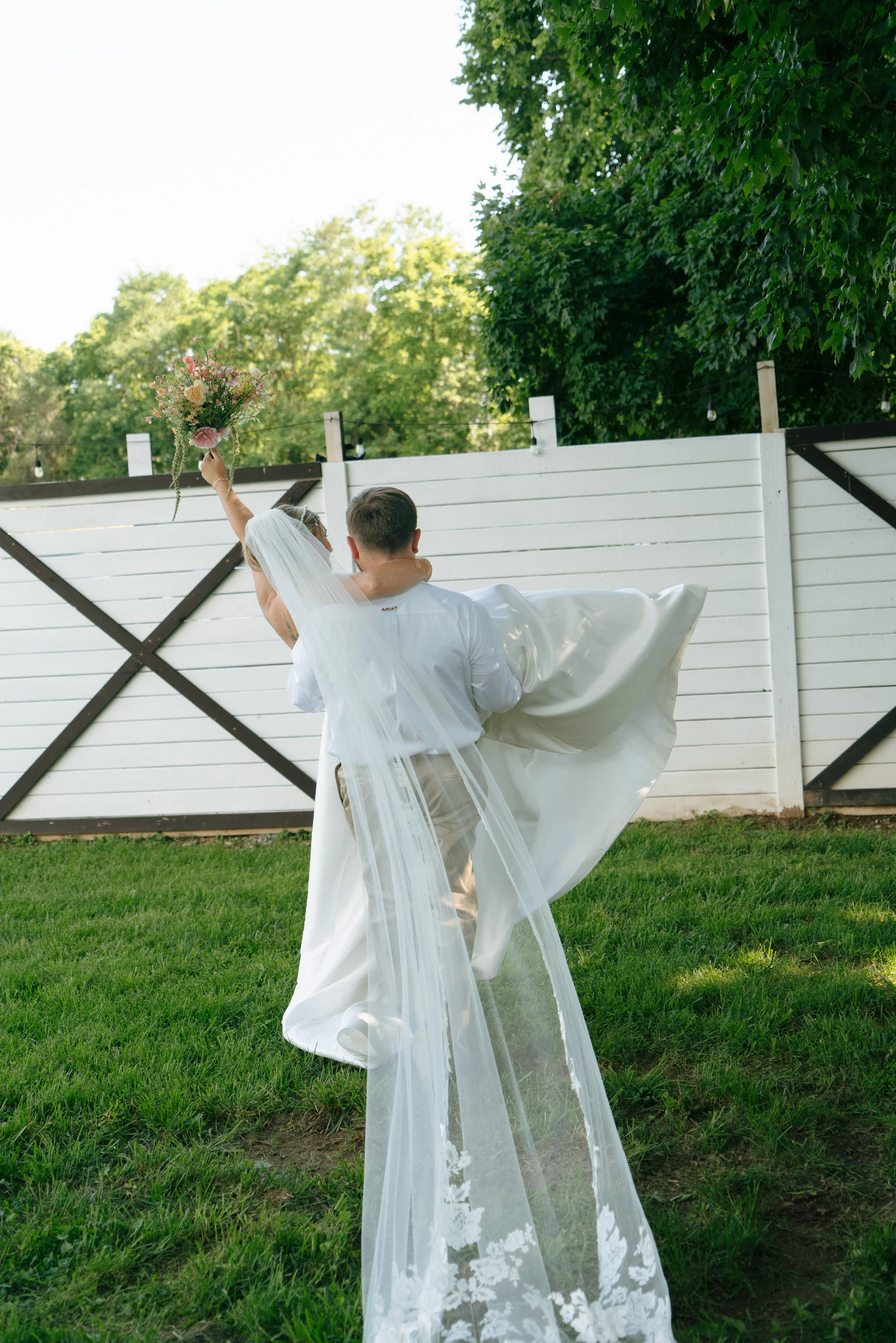 A newlywed couple is outdoors in a grassy backyard with a white fence and green trees in the background. The bride, wearing a wedding veil and gown, is being carried by the groom. She is holding a bouquet of flowers high in the air.
