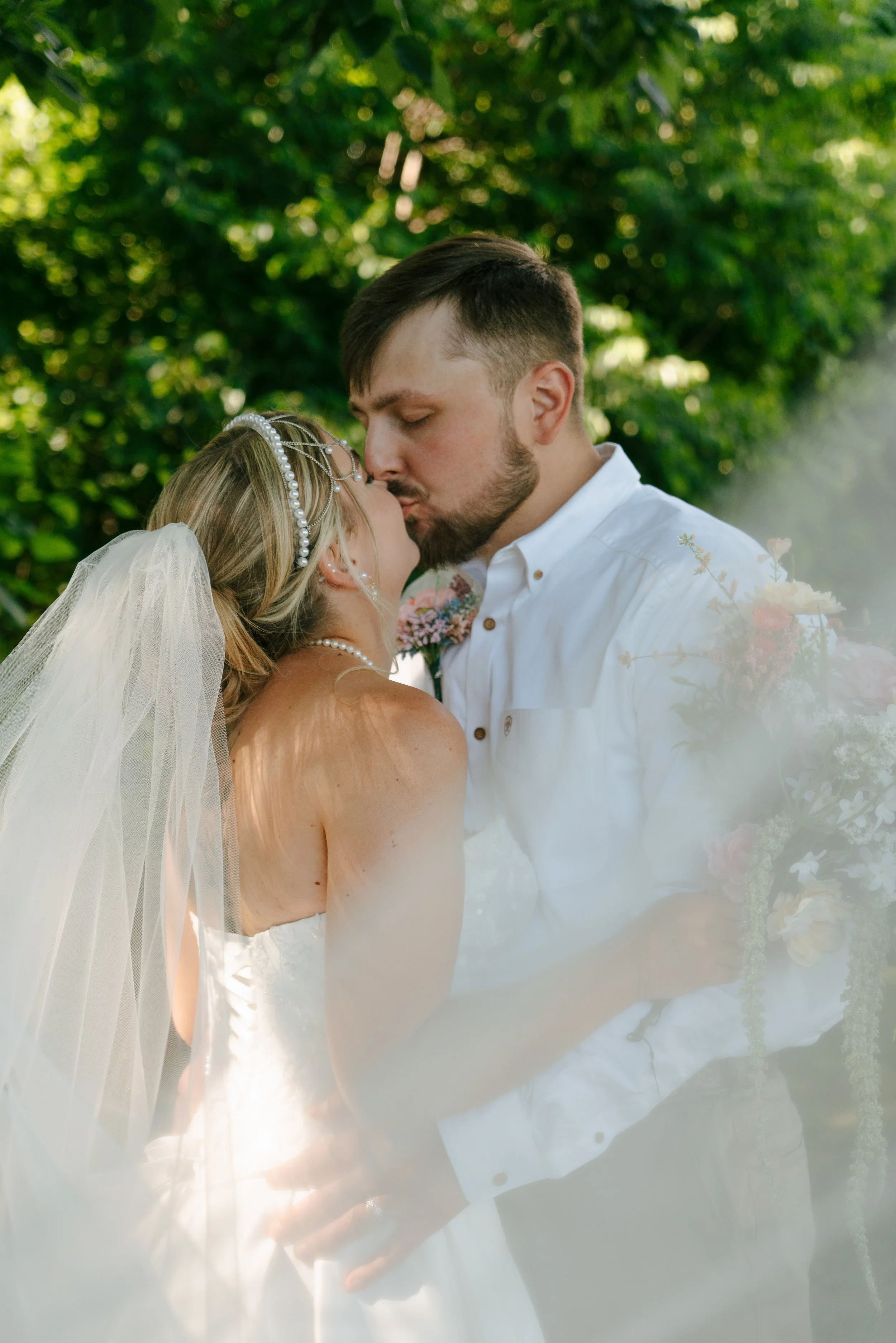 A man and a woman kissing outdoors, with greenery in the background. The woman has a veil and pearl headband, and the man is wearing a white shirt. The woman is holding the man's hand.