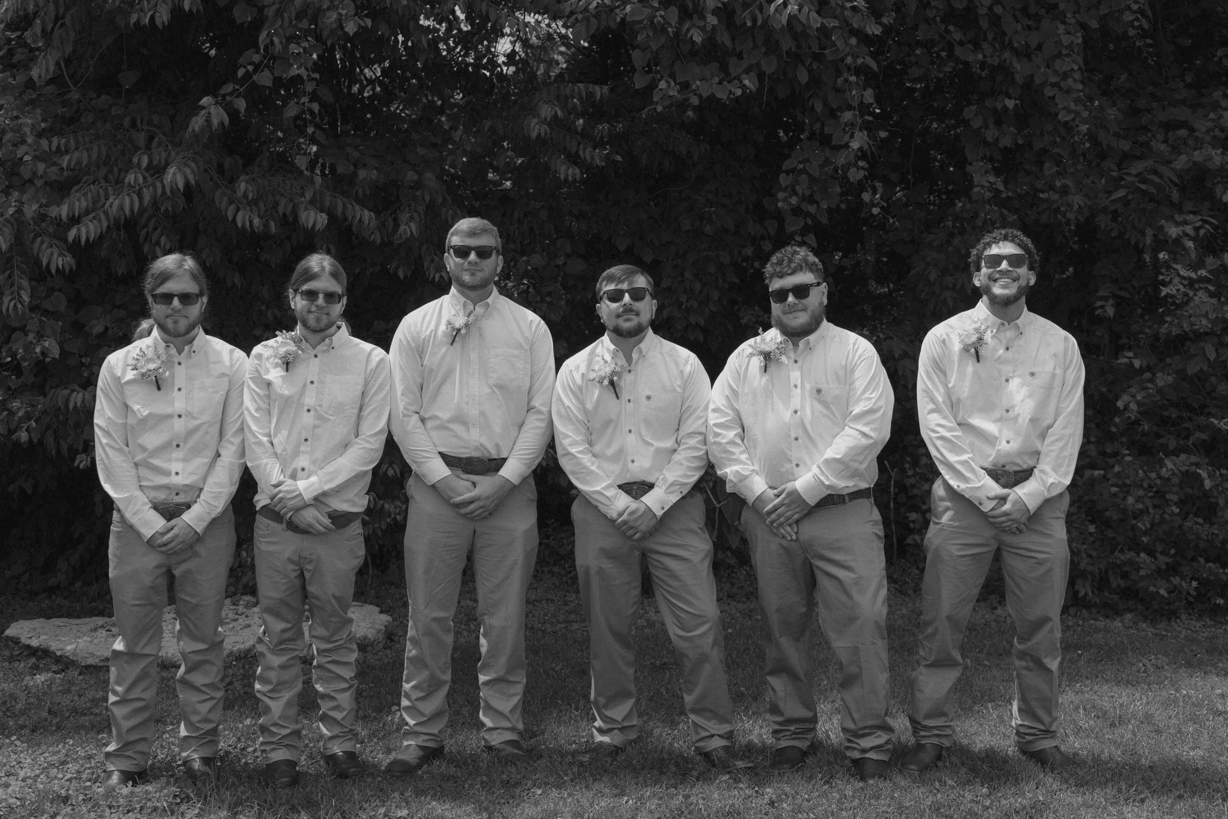 A group of six men wearing white shirts, khaki pants, sunglasses, and boutonnières, standing outdoors in front of leafy trees.
