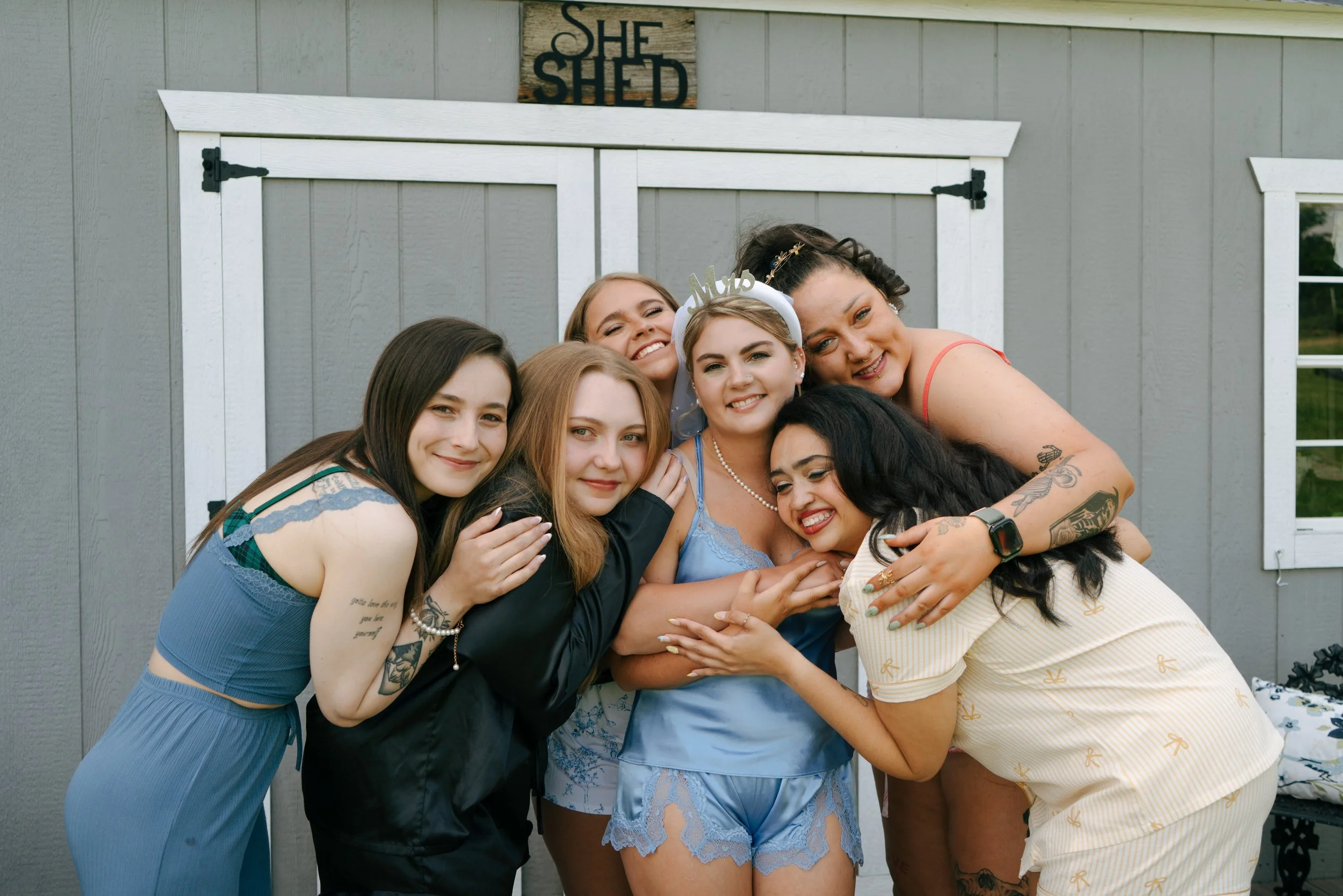 A group of six women hugging and smiling in front of a gray shed with a wooden sign that says "SHE SHED."
