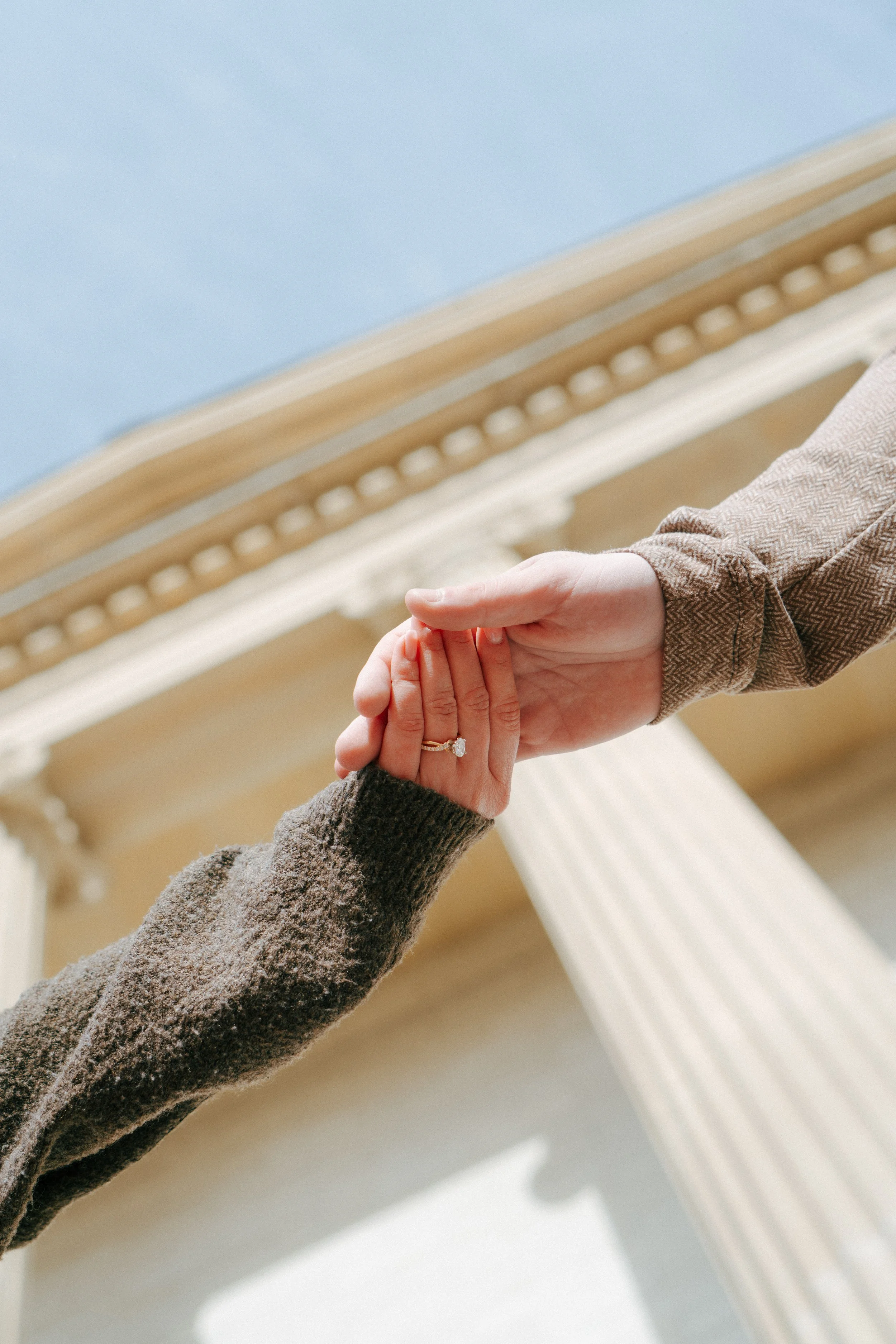 Two people are holding hands, with one person wearing a ring on their finger. The photo is taken indoors near a ceiling with decorative molding.