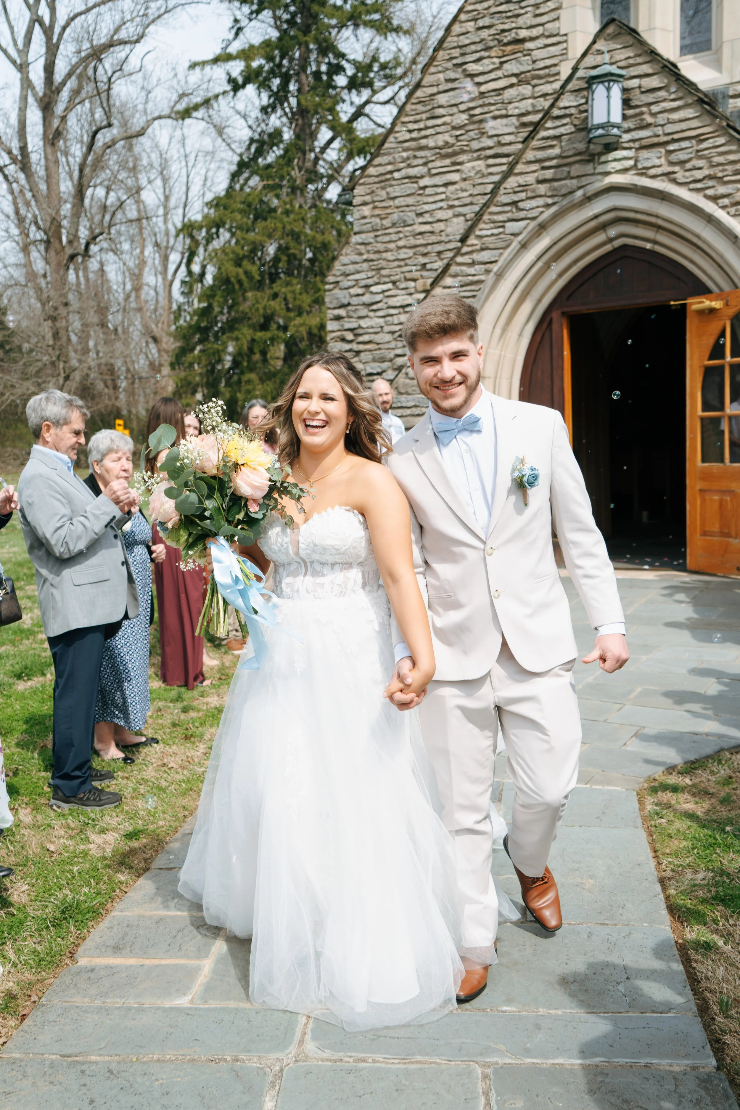 A bride and groom smiling and holding hands outside a church after their wedding, with guests in the background.