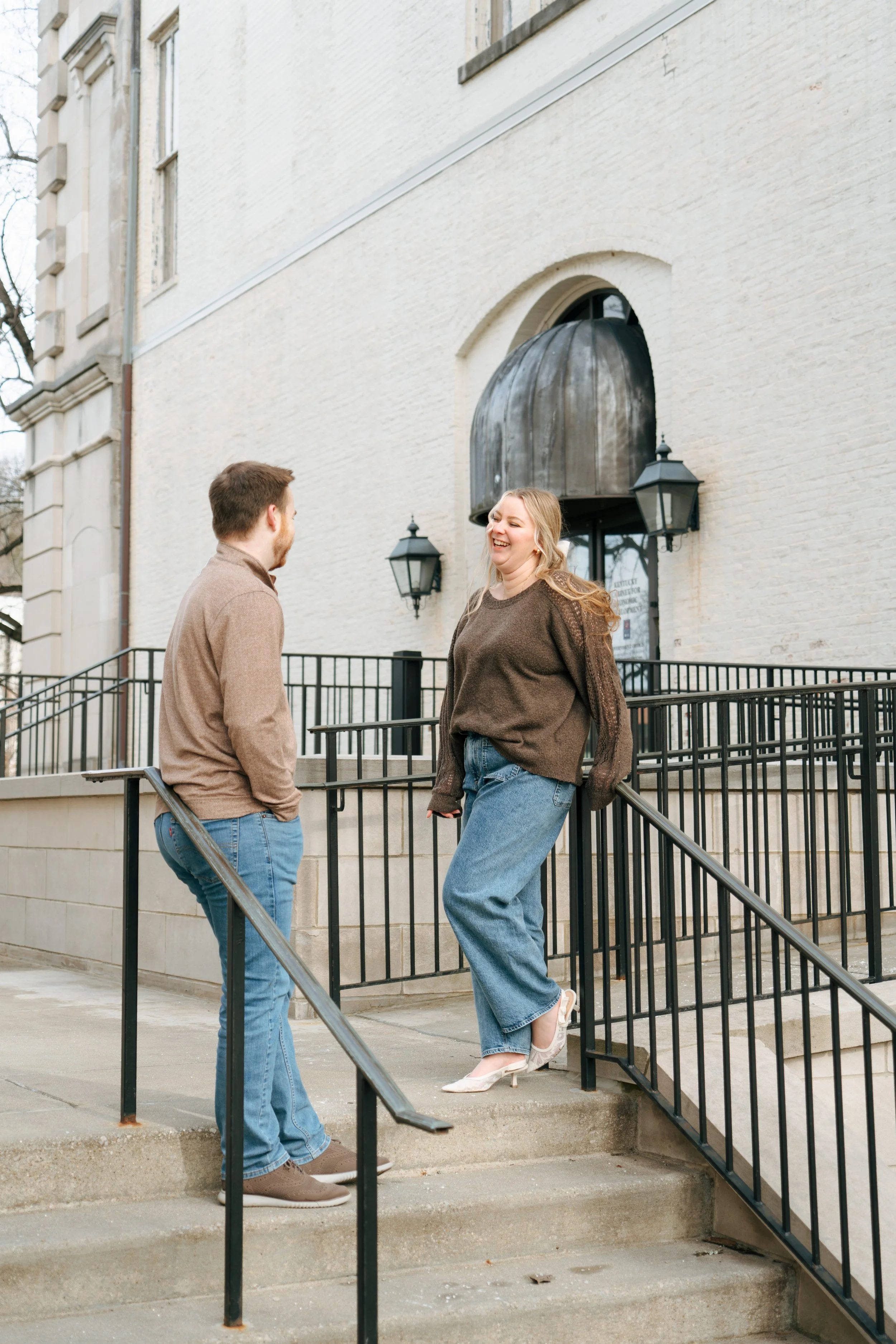 A woman wearing a brown sweater, wide-leg jeans, and white high heels is smiling and laughing while talking to a man in a tan jacket and blue jeans outside a white brick building with black lamp fixtures. They are standing on concrete steps with a bl
