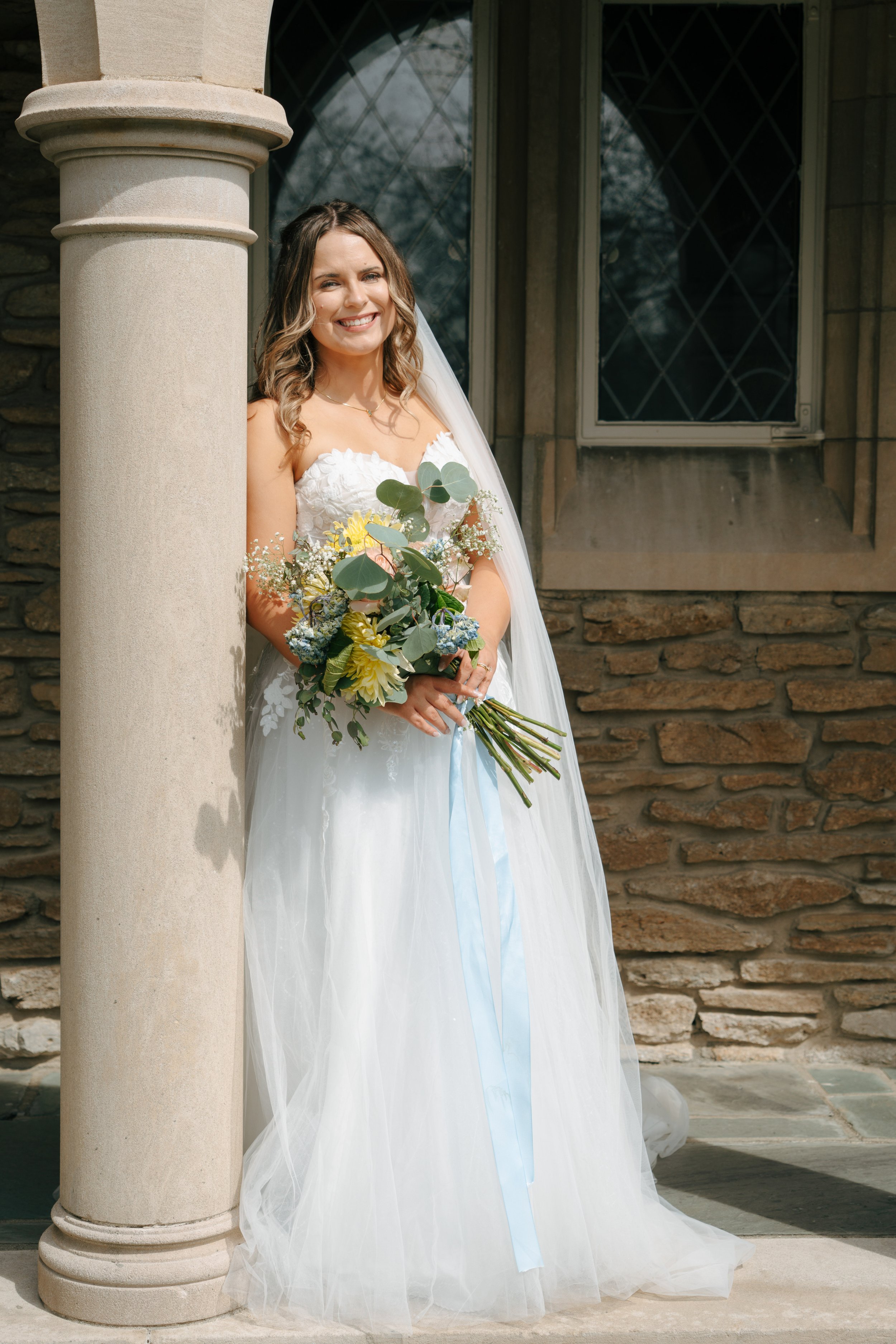 A smiling bride with wavy brown hair, wearing a white wedding dress and veil, standing outside near a stone column and a window with diamond-shaped panes, holding a bouquet of yellow, pink, white, and blue flowers with greenery.