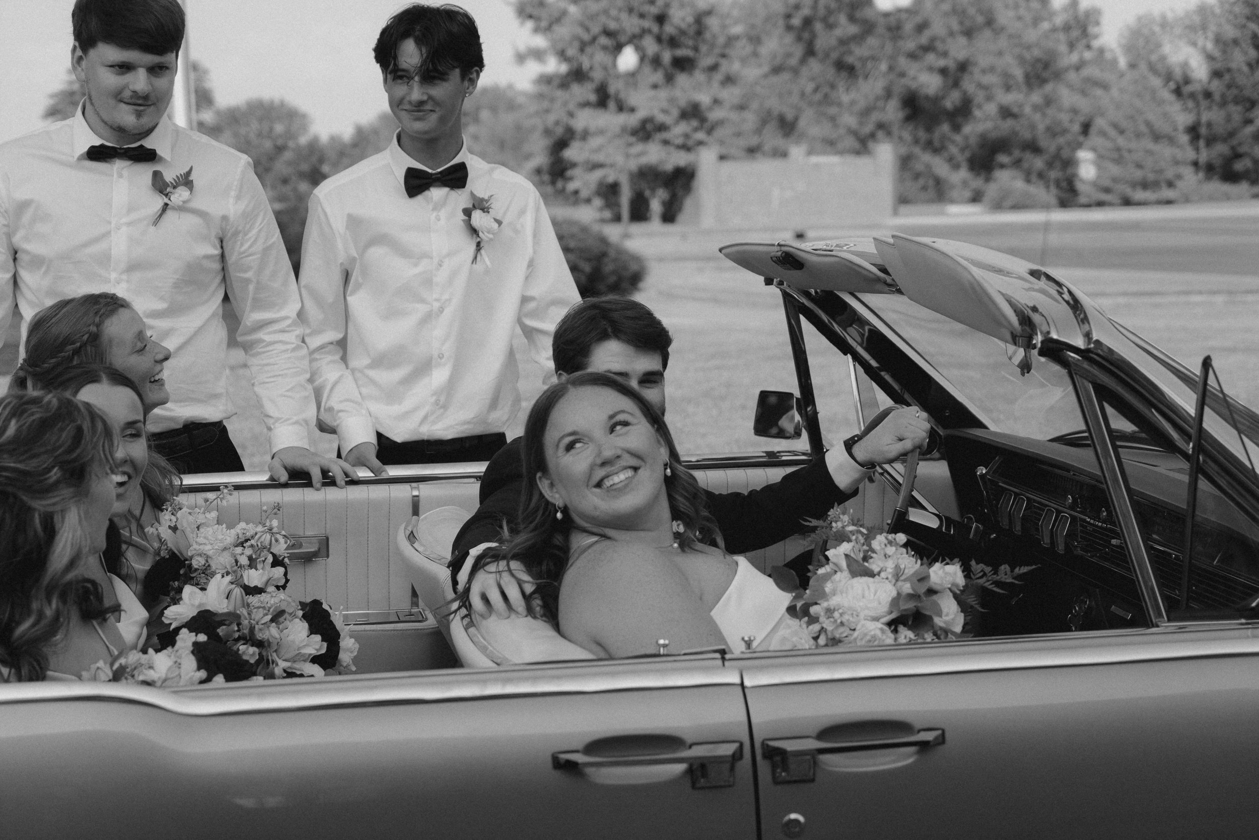 Black and white photo of a wedding party in a convertible car, with two men dressed in tuxedos with bow ties and boutonnières standing behind the women, who are seated in the car. The women are smiling and wearing dresses with floral accessories. The