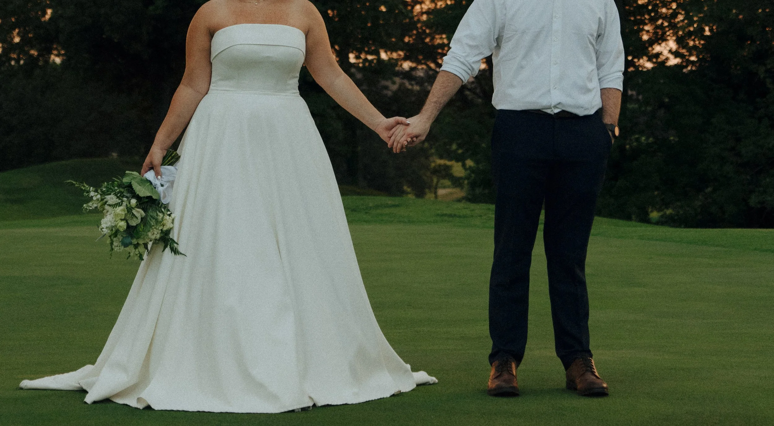 A bride and groom holding hands outdoors during twilight. The bride wears a white strapless wedding dress and holds a bouquet of white and green flowers. The groom is dressed in a white shirt, dark pants, and brown shoes. The background features tree