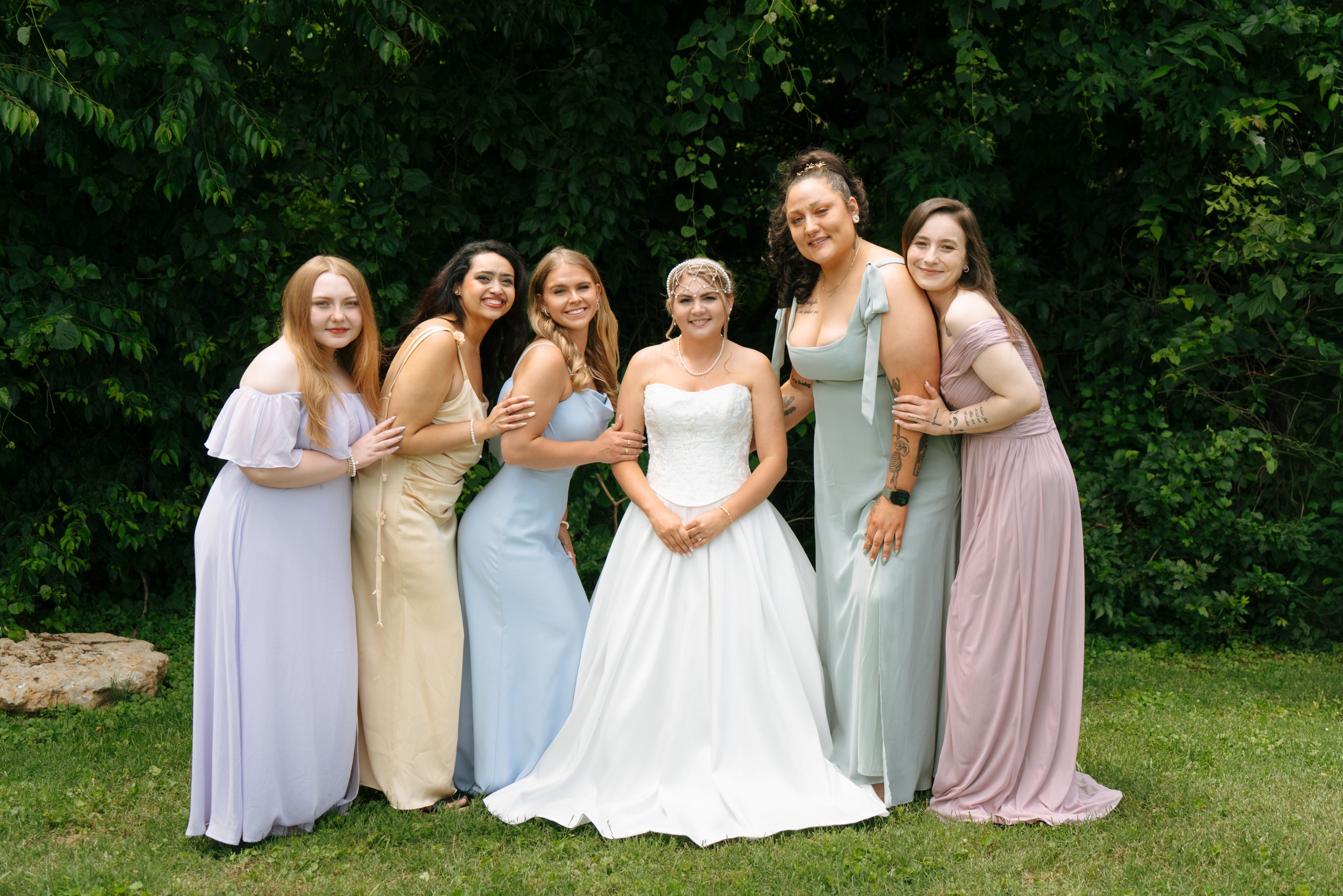 Group of seven women, one in a wedding dress, outdoors in front of green leafy background, smiling and posing for a photo.