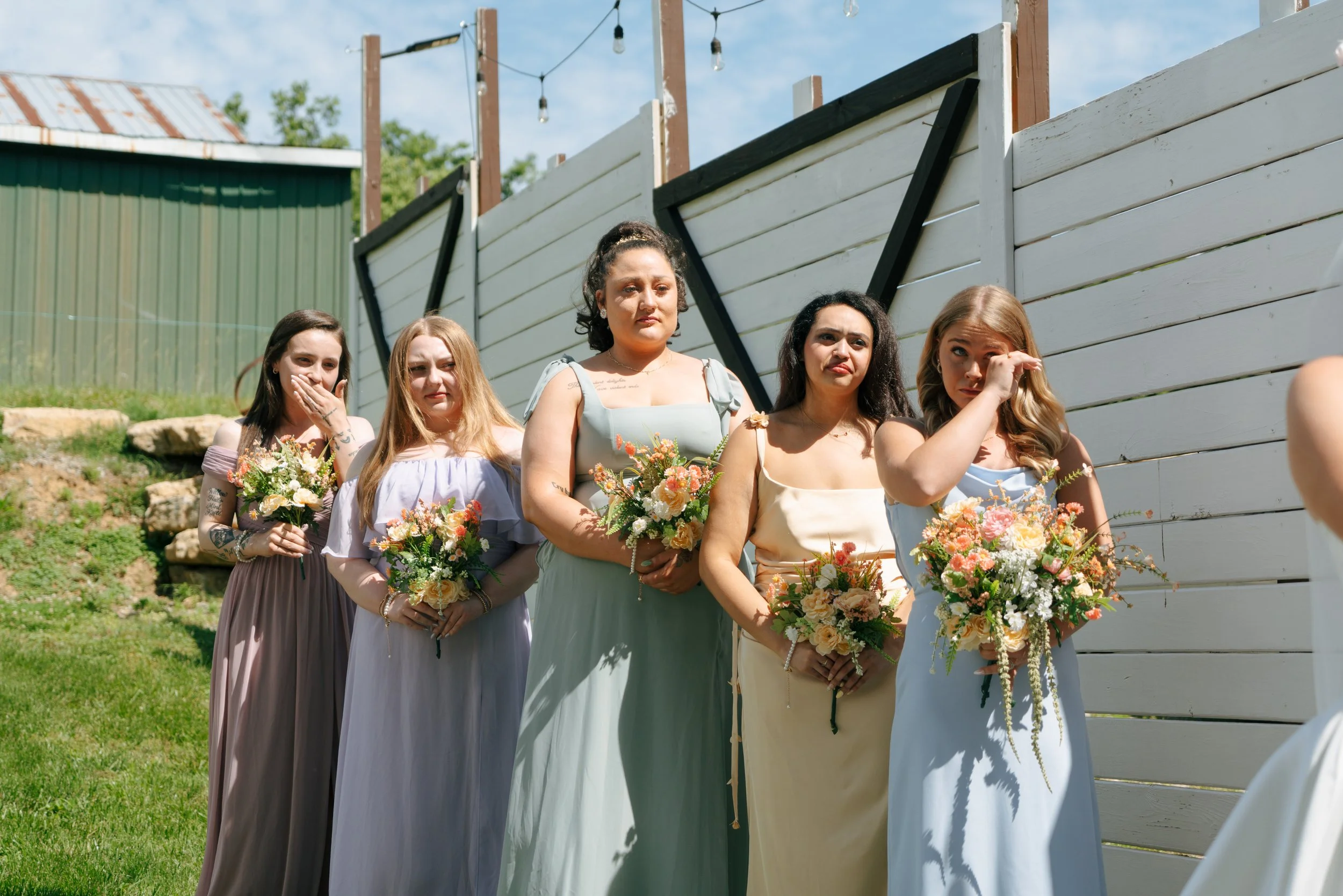 Group of five women standing outdoors during a wedding ceremony, holding bouquets of flowers, in front of a white wooden fence with black accents and string lights overhead.