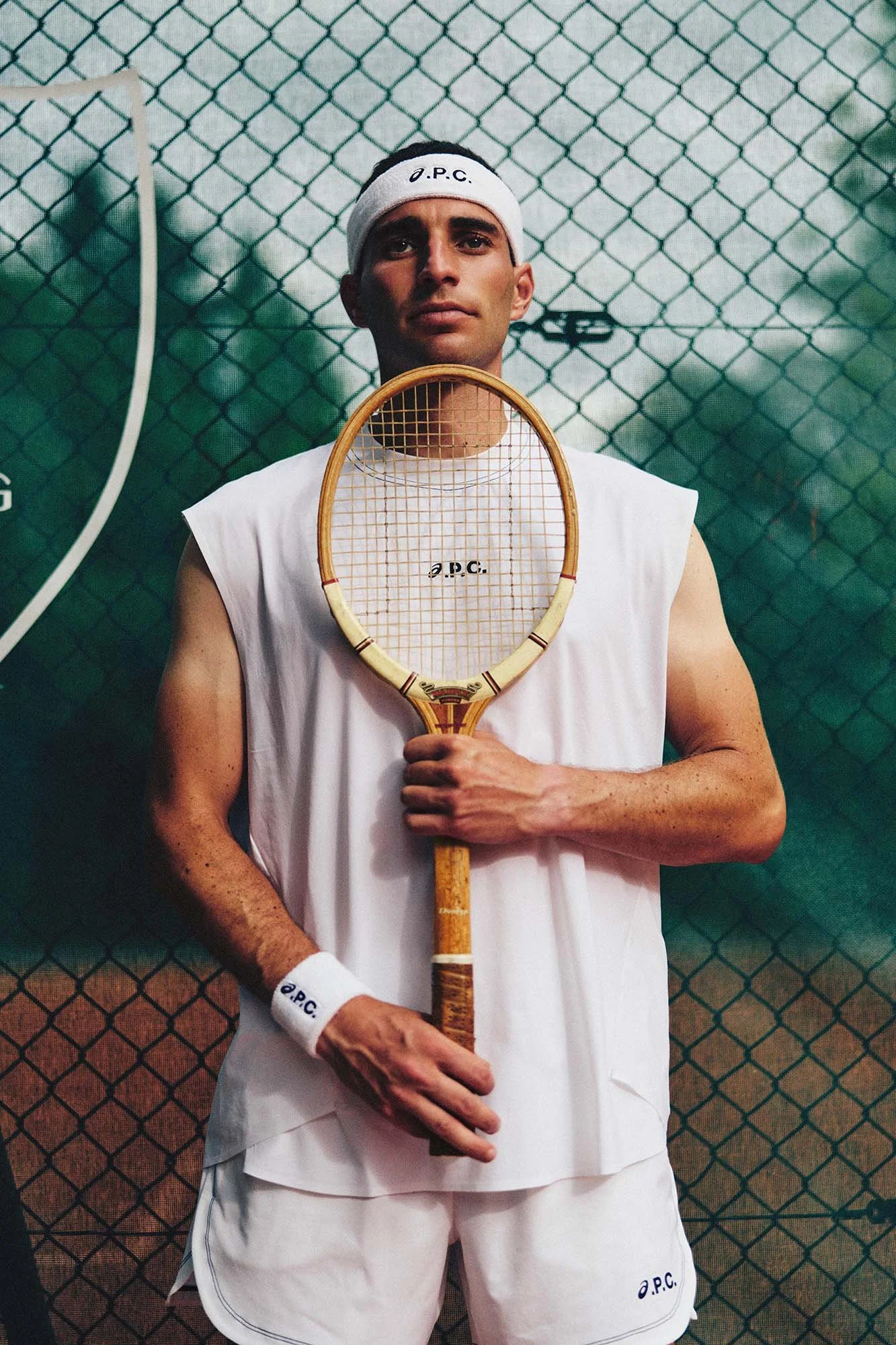Male model in white A.P.C. tennis apparel holding a vintage wooden tennis racket in front of a green chain-link fence.