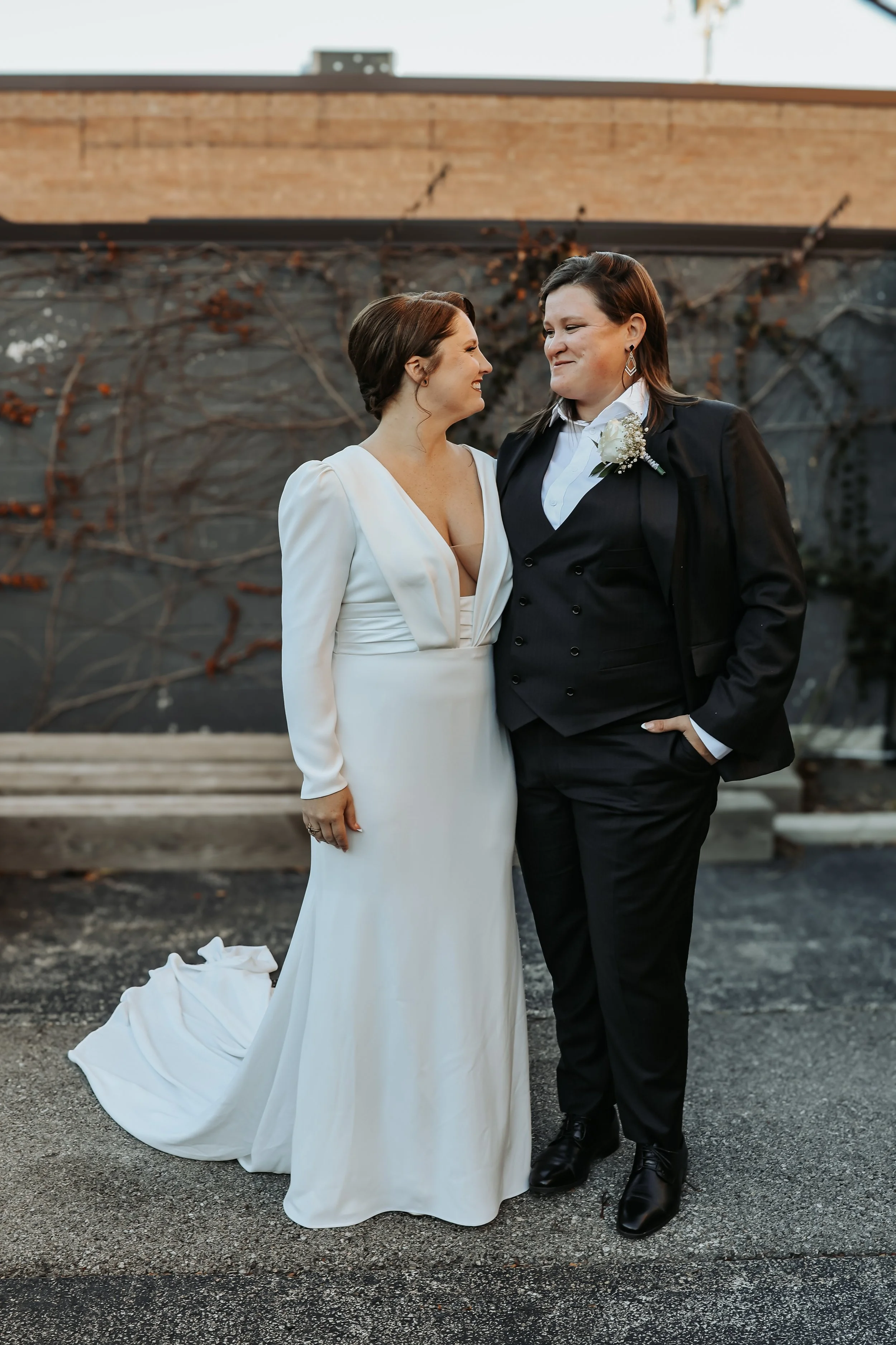 A couple, one in a white wedding dress and the other in a black suit, standing close together and smiling at each other outdoors at Eris Brewery and Cider House in Chicago, IL.