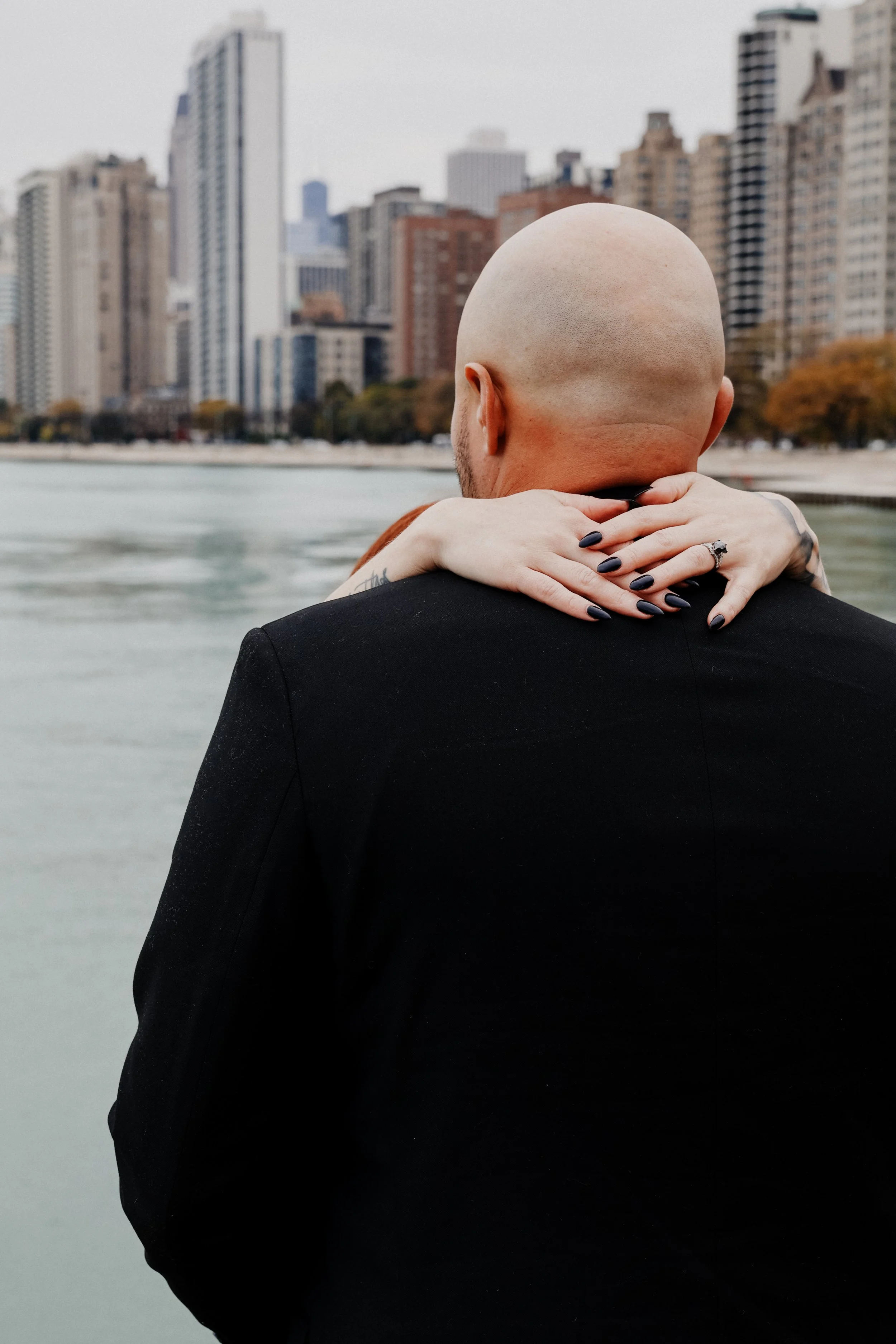 A bride's hands are around her groom's neck on a lakefront path with Chicago skyline in the background.