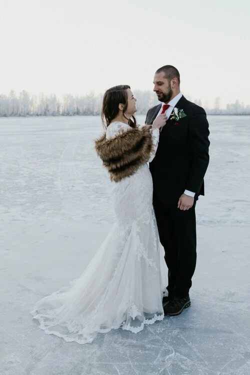 A bride and groom standing on an ice-covered lake during winter. The bride is wearing a white lace wedding gown and a fur stole, while the groom is in a black suit with a white shirt, red tie, and boutonniere. They are looking at each other affectionately.