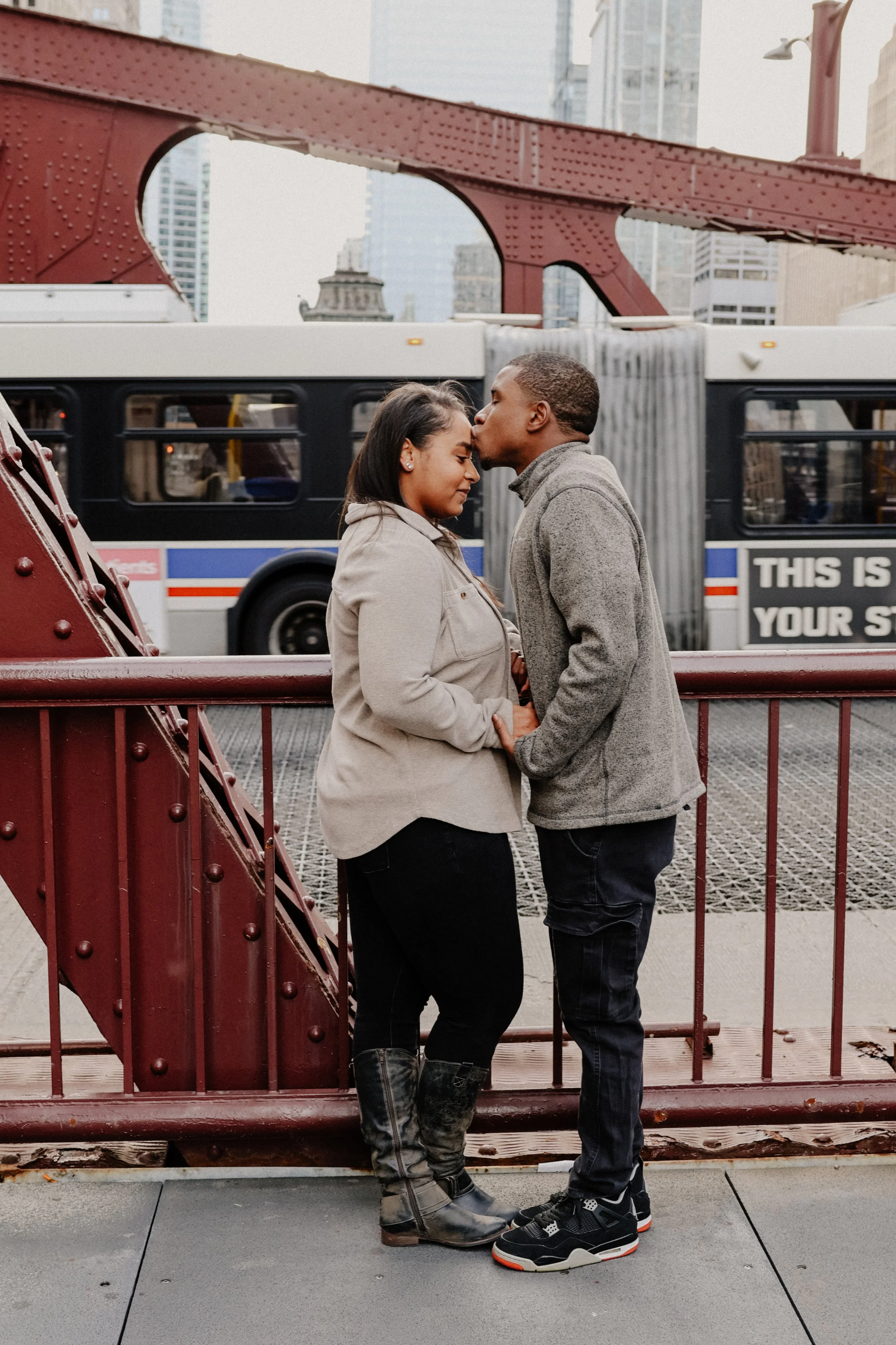 A groom-to-be kisses his bride's forehead on a bridge in downtown Chicago as a bus passes by in the background.