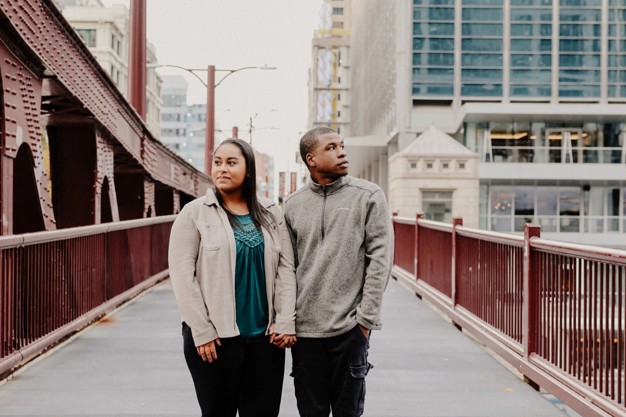 Two people stand on a bridge on the Riverwalk in Chicago looking away from each other as they pose for engagement photos.