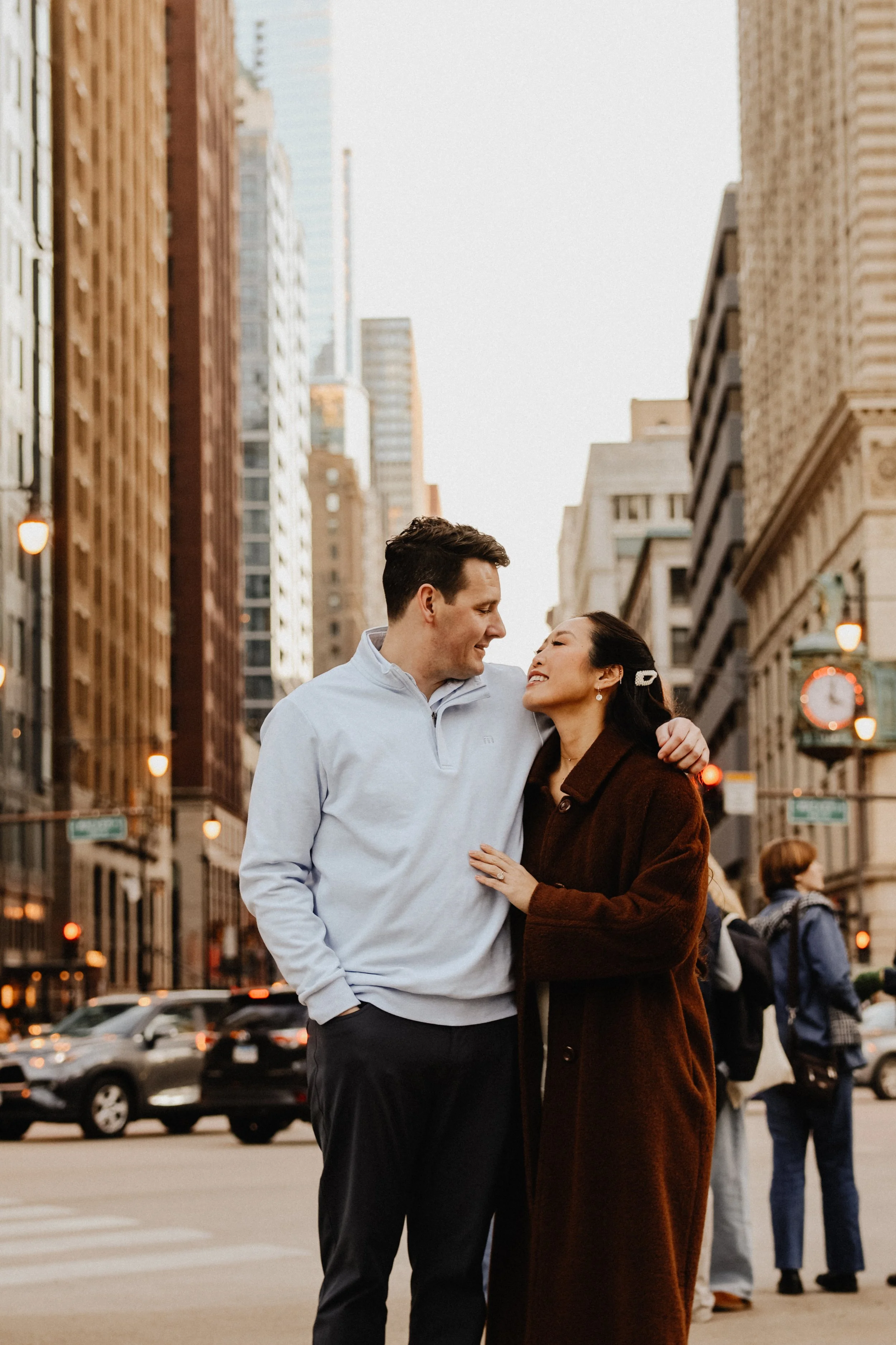 A couple stands over a manhole cover in downtown Chicago at an intersection posing for engagement photos with the skyline in the background.