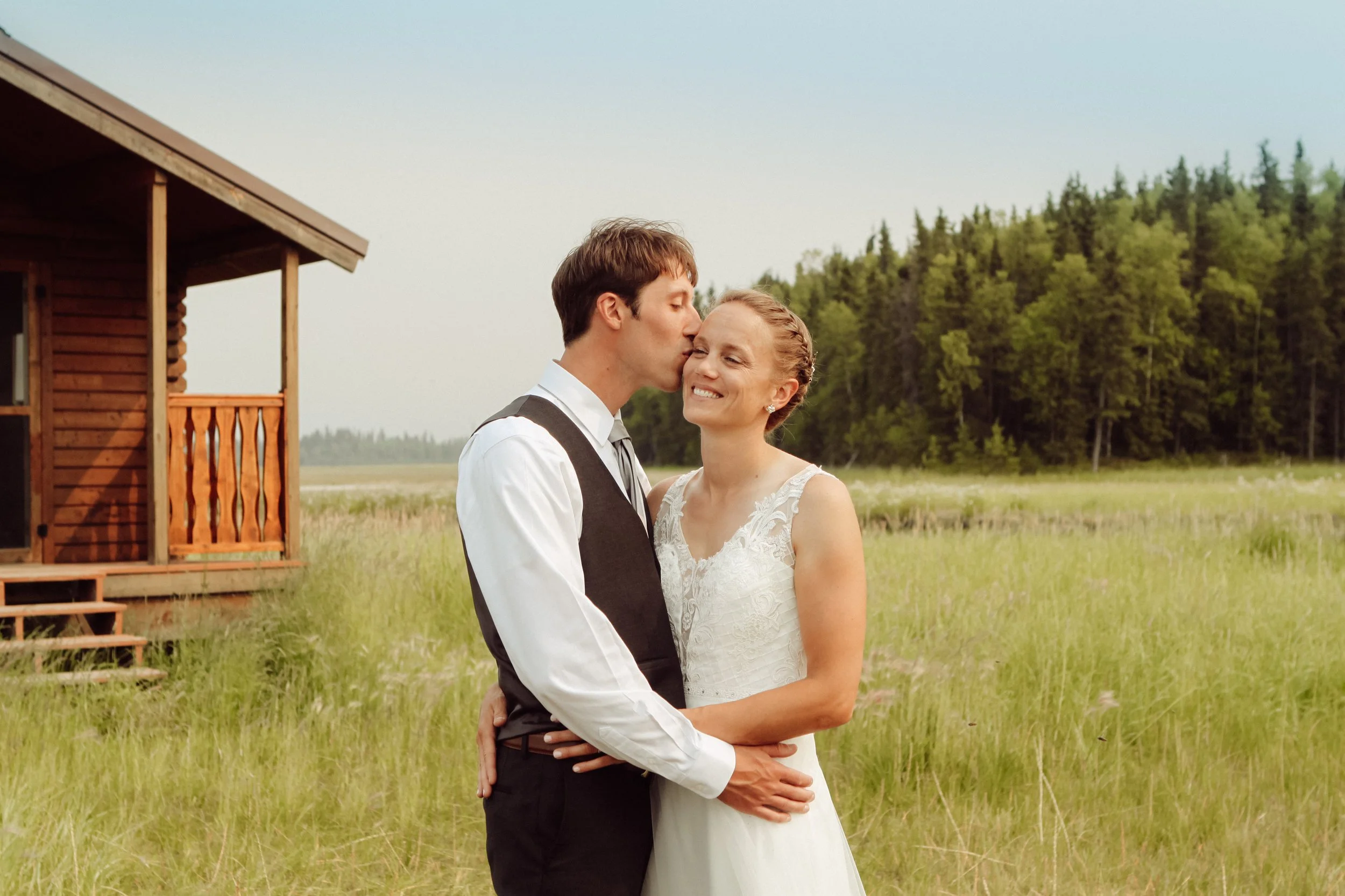 A bride and groom standing outside on a grassy field near a wooden cabin, with the groom kissing the bride on the forehead, both smiling happily.