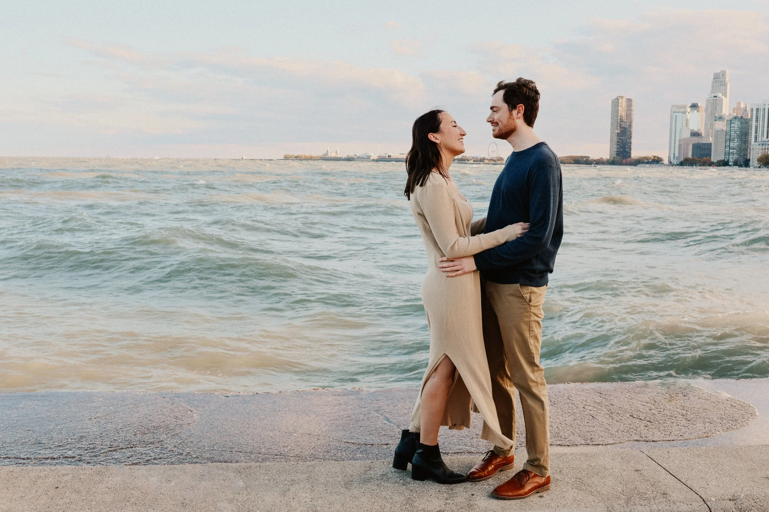 An engaged couple standing on North Avenue Beach in Chicago, IL on the lakefront holding each other and smiling at each other.
