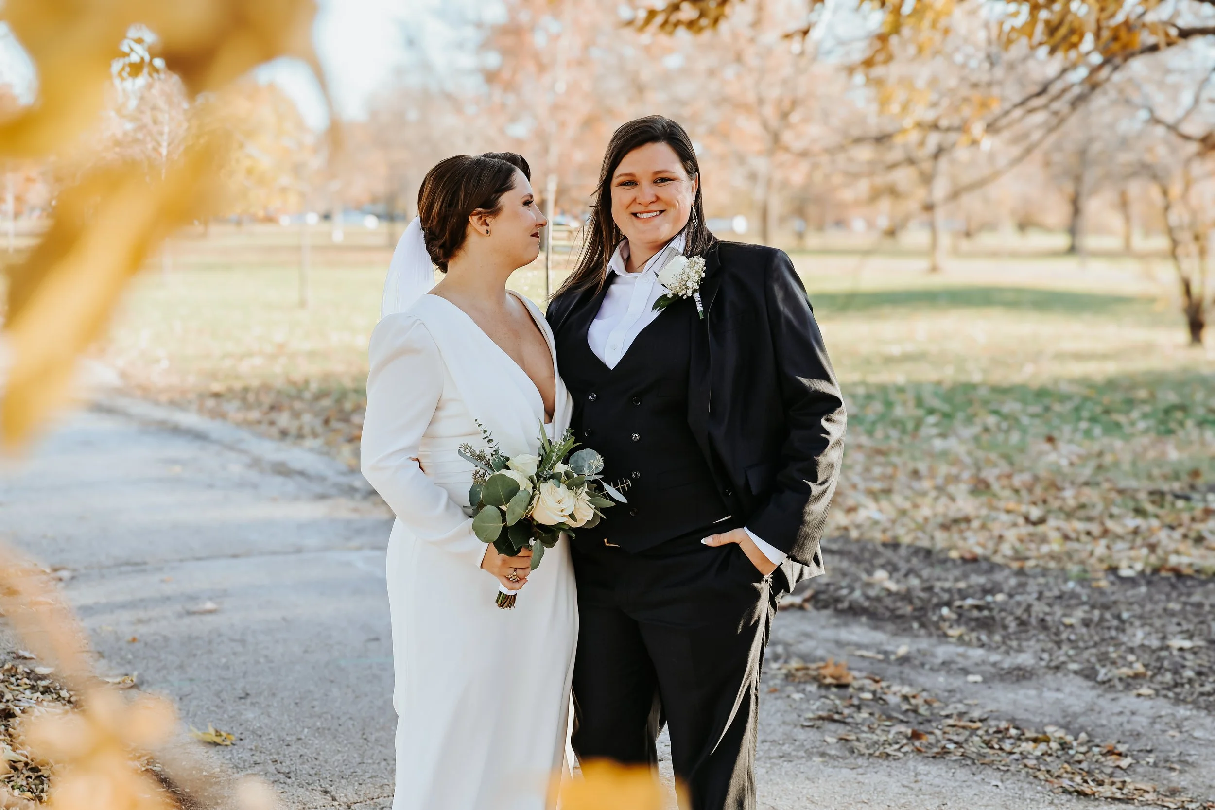 Two brides stand together in beautiful Humboldt Park in Chicago in the fall with a wedding gown and suit. They stand on a path bordered by foliage.