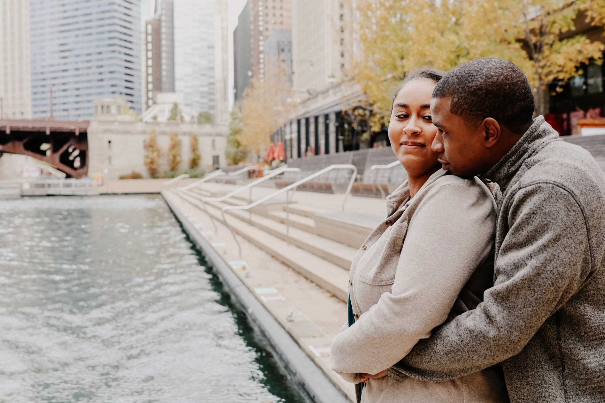 A groom hugs his bride from behind as she looks over her shoulder at him, posing for engagement photos in Chicago.